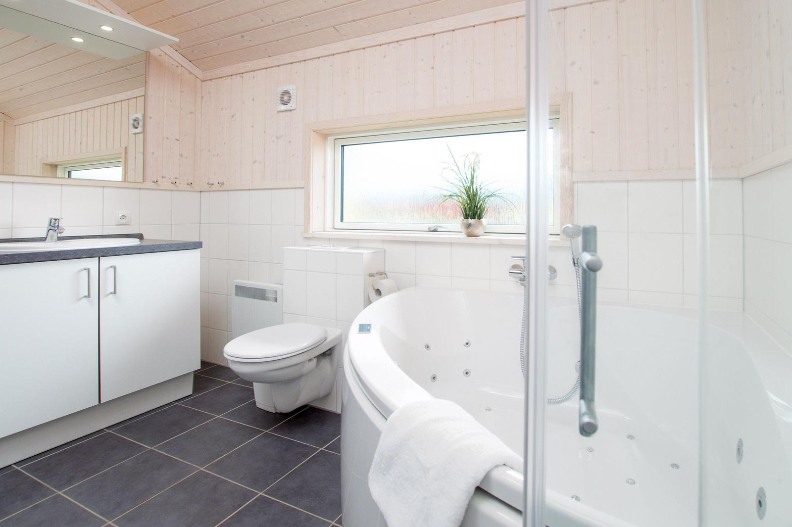 Badezimmer mit Badewanne, Toilette und Waschbecken. Holzwände und Fenster mit Pflanze.