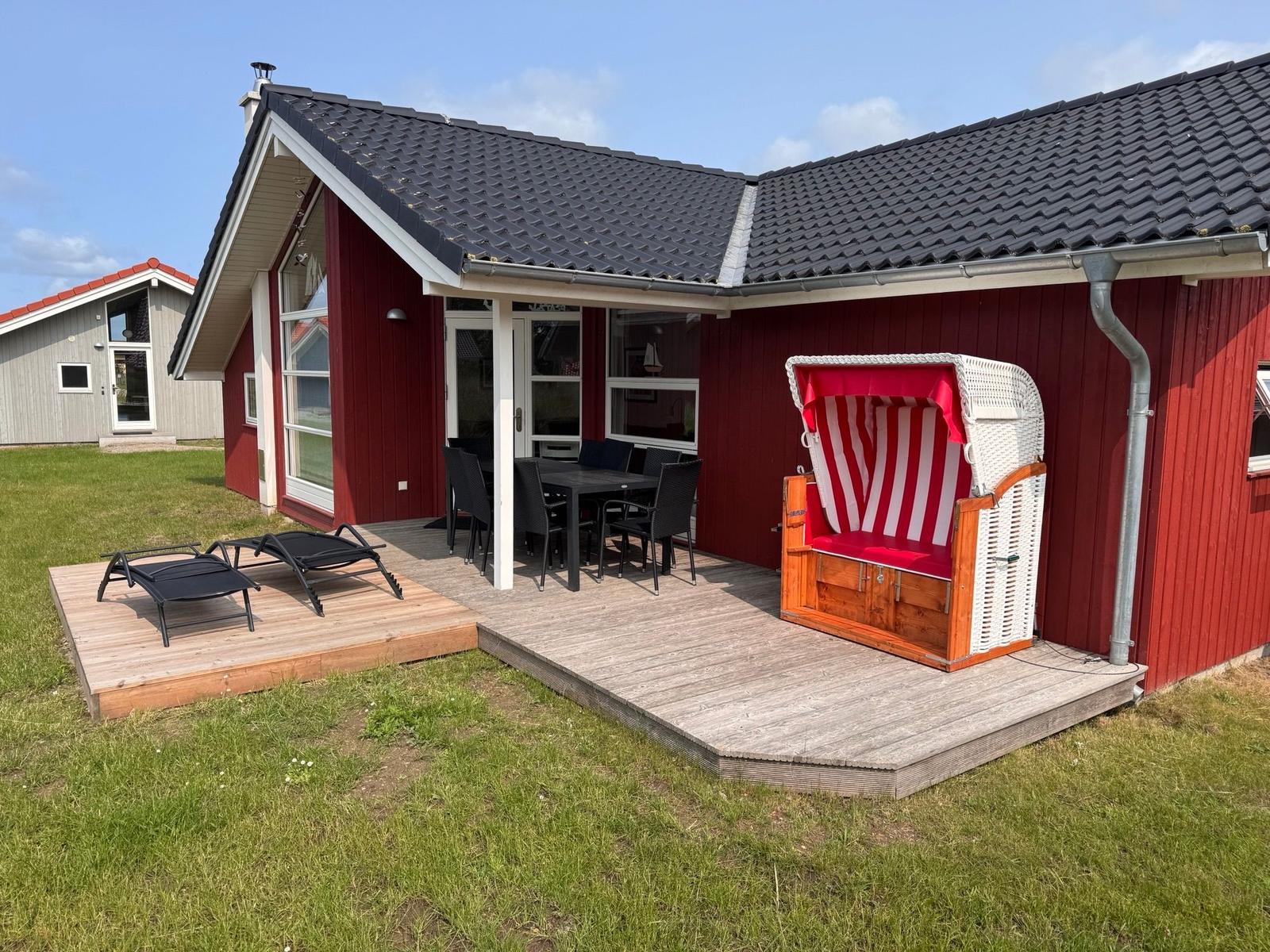 Rotes Ferienhaus mit Terrasse, Liegen und Strandkorb. Grünfläche und klarer Himmel.
