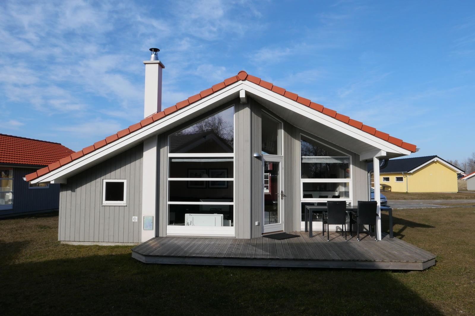 Moderne Ferienhaus mit Holzterrasse, großen Fenstern und rotem Dachziegel.