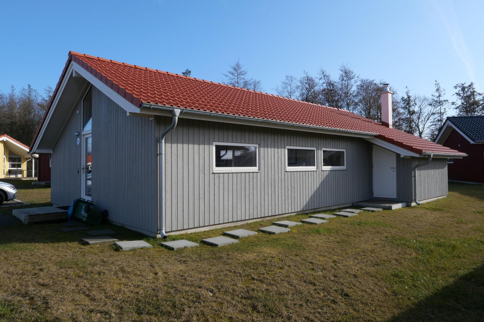 Ein graues Holzhaus mit rotem Dach und weißen Fenstern. Grasfläche und Steine zum Eingang.