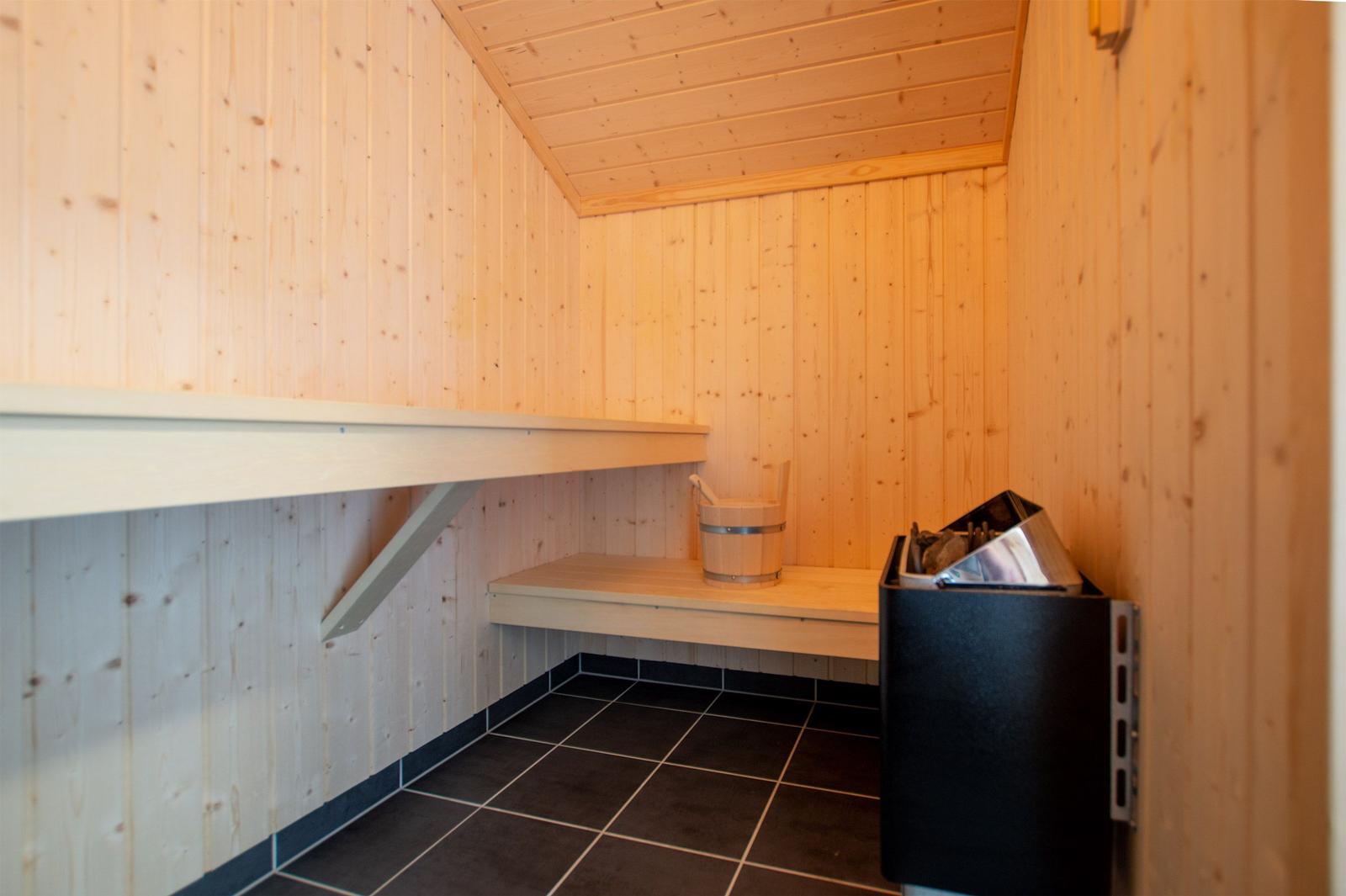 Sauna mit Holzwand, Sitzbänken und Holzmulde. Auf dem Boden liegen schwarze Fliesen.