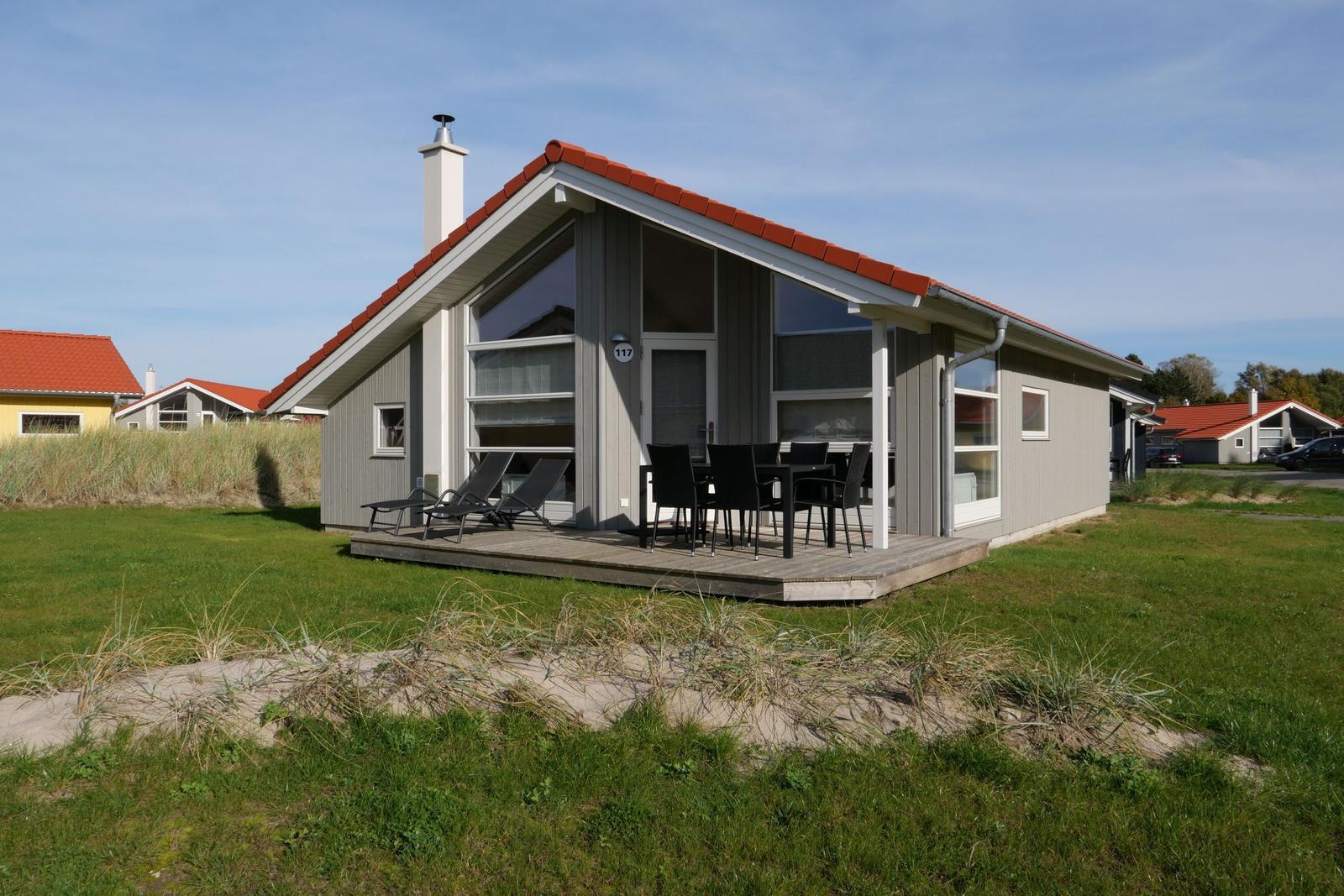Moderne Ferienhaus mit Terrasse, Tisch und Stühlen, umgeben von Gras und Sanddünen.