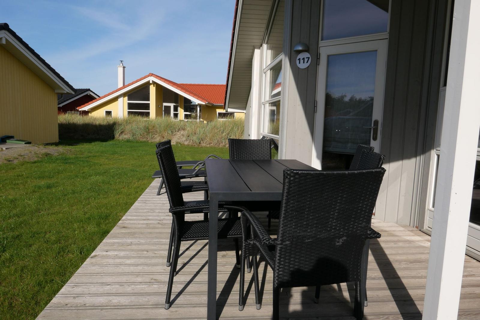Terrasse mit Tisch und Stühlen vor Haus 117. Grasfläche und Nachbarhäuser im Hintergrund.