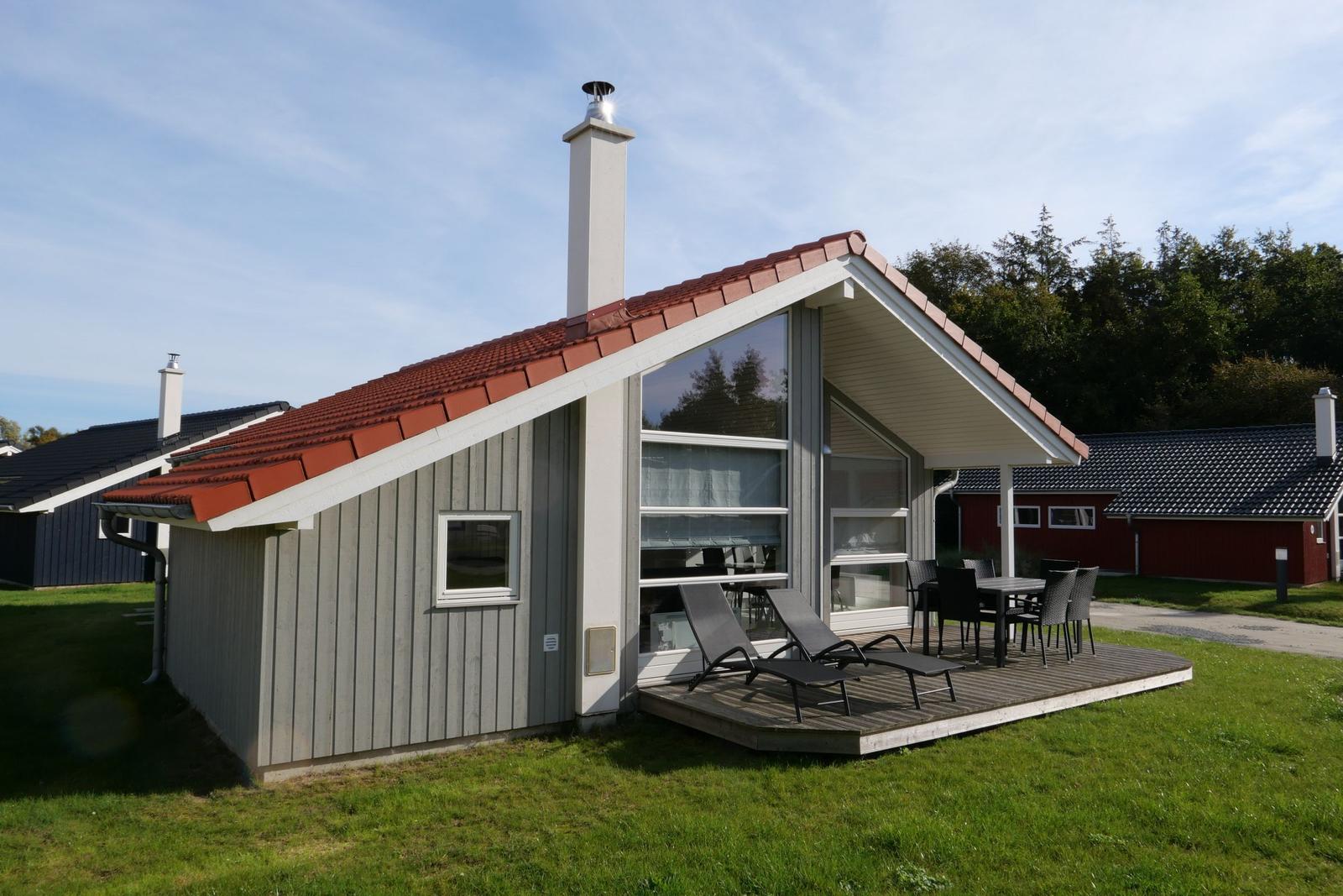 Moderne Holzhütte mit Terrasse, Sonnenliegen und Tisch. Roter Dachfirst und Schornstein.