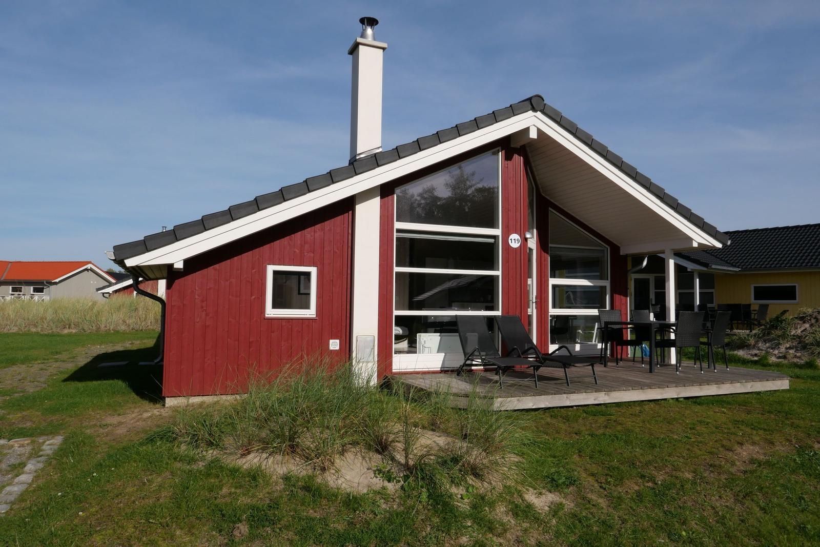 Rotes Holzhaus mit Terrasse, Sonnenliegen und Tisch. Rauchfangschornstein und große Fenster.
