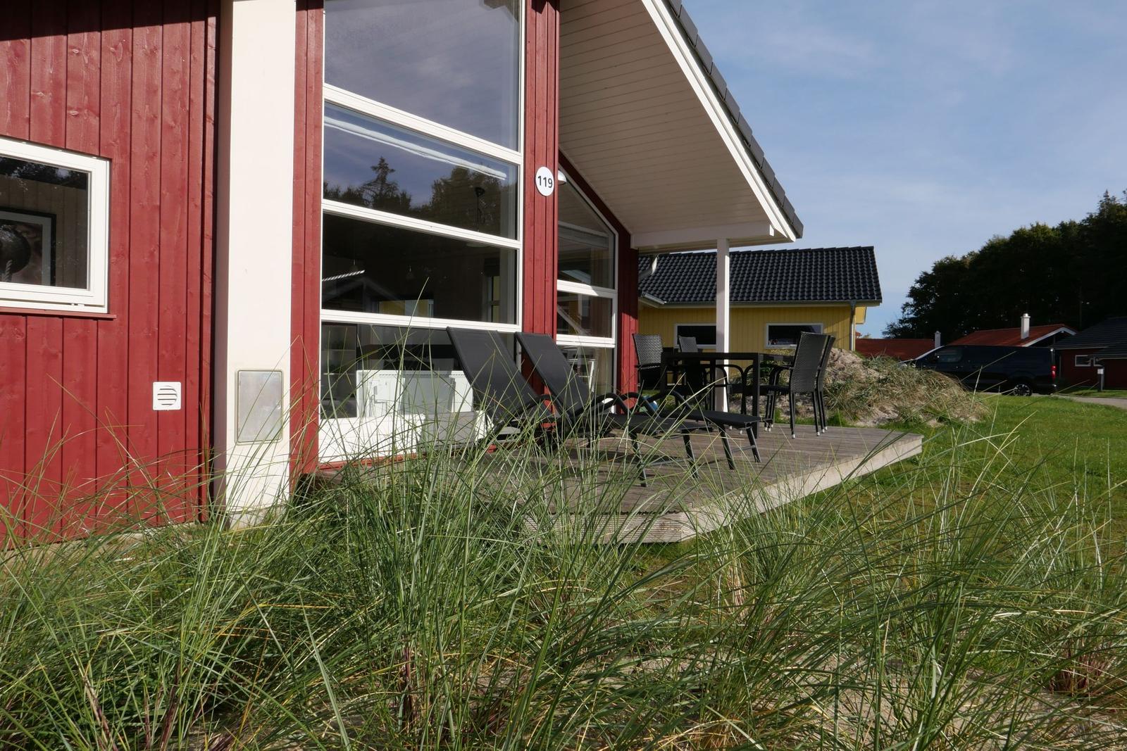 Rotes Ferienhaus mit Terrasse, Liegen und Tisch. Gras im Vordergrund.