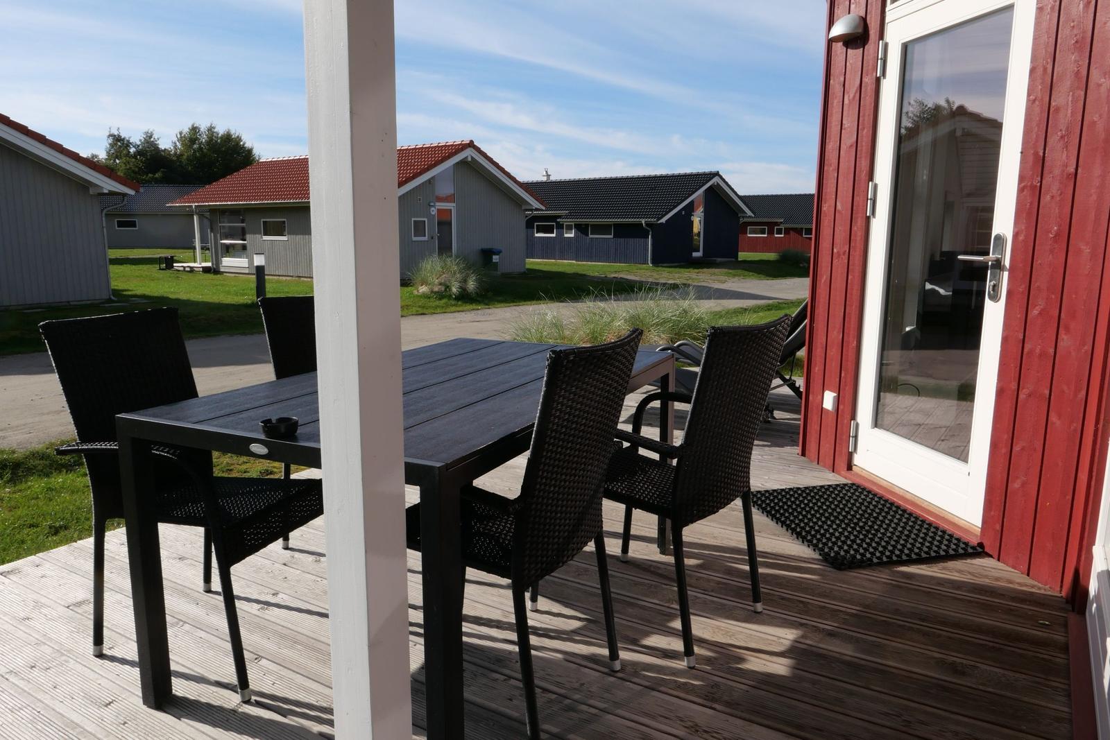 Terrasse mit Tisch und Stühlen vor einem roten Haus. Im Hintergrund sind andere Häuser und Grünflächen sichtbar.