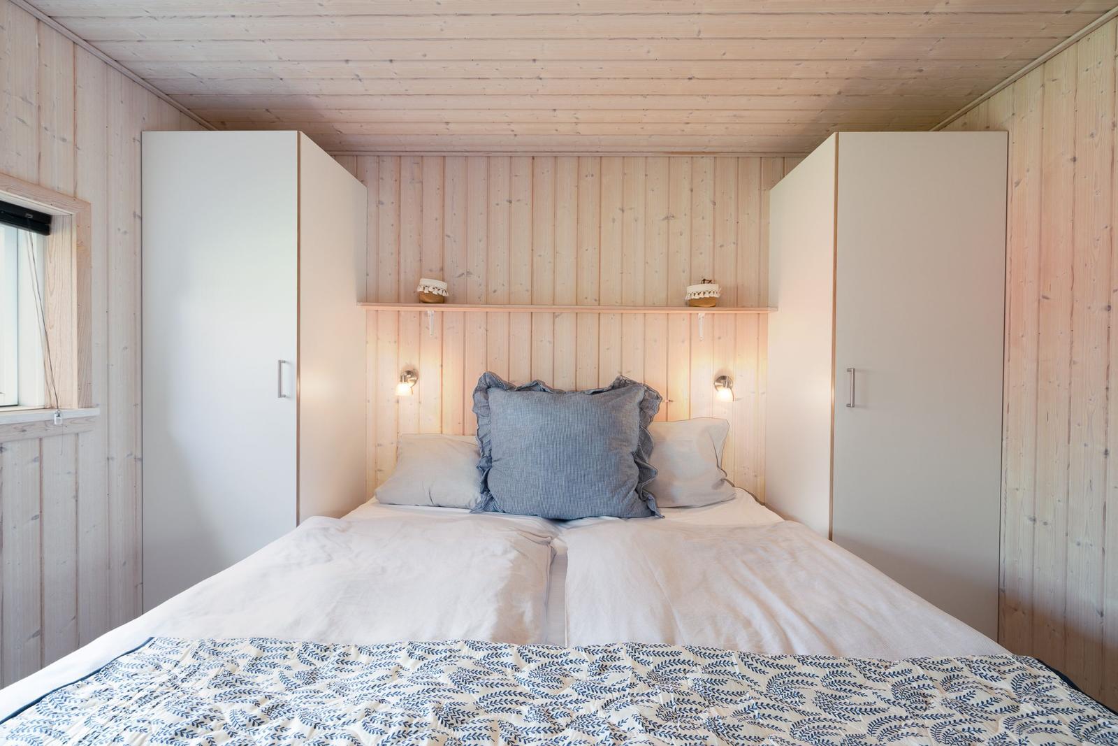 Schlafzimmer mit Bett, weißen Schränken und Holzwandverkleidung. Kopfteil mit Nachttischbeleuchtung.