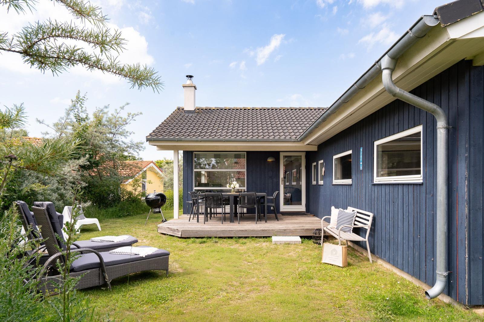 Ein blaues Haus mit Terrasse, Gartenliegen und Grill. Hintergrund: Bäume und Himmel.