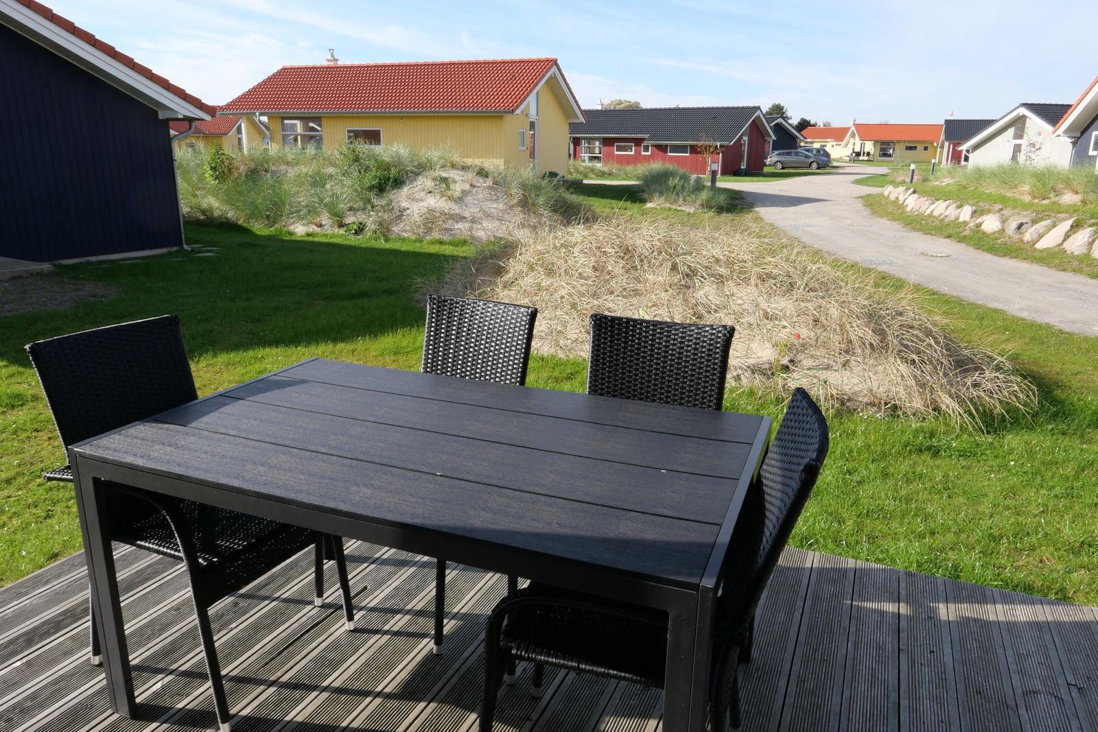Schwarzer Tisch mit vier Stühlen auf Terrasse. Gras und Häuser im Hintergrund.