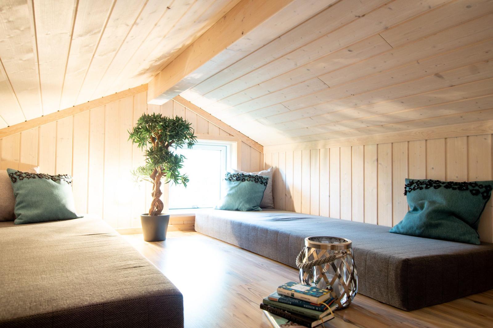 Zimmer mit Holzwänden, Fenster, Pflanze und Sitzbänken mit Kissen.