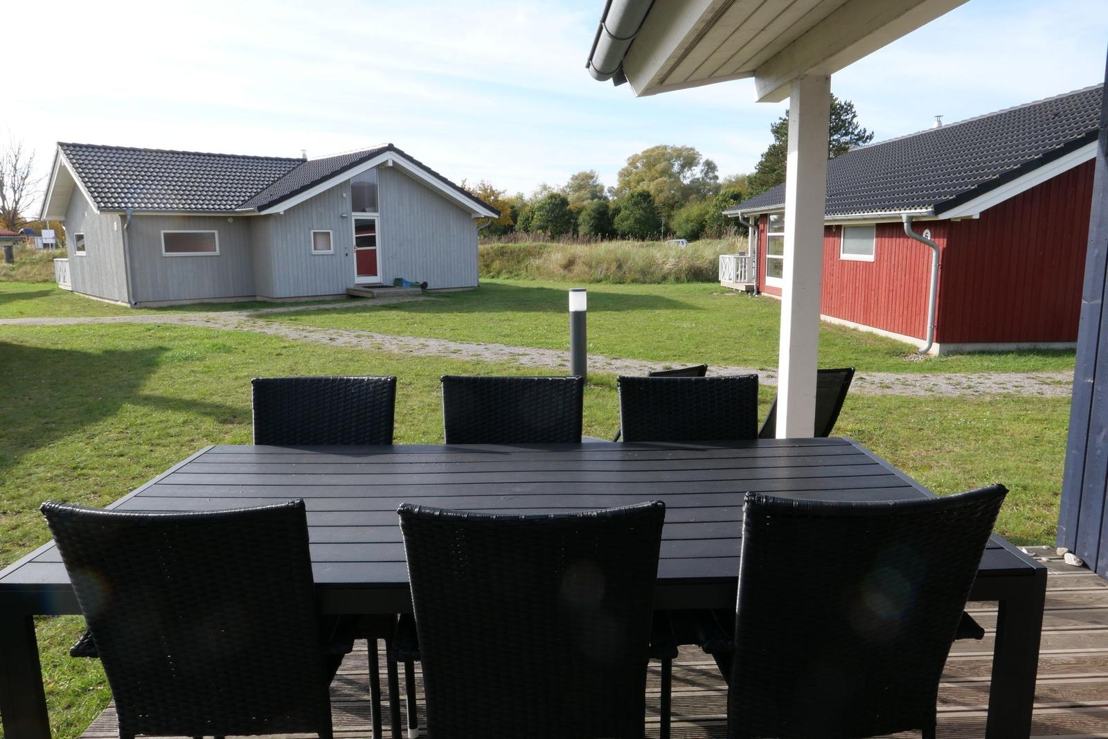 Terrasse mit Tisch und Stühlen. Im Hintergrund sind Häuser und Grasland zu sehen.