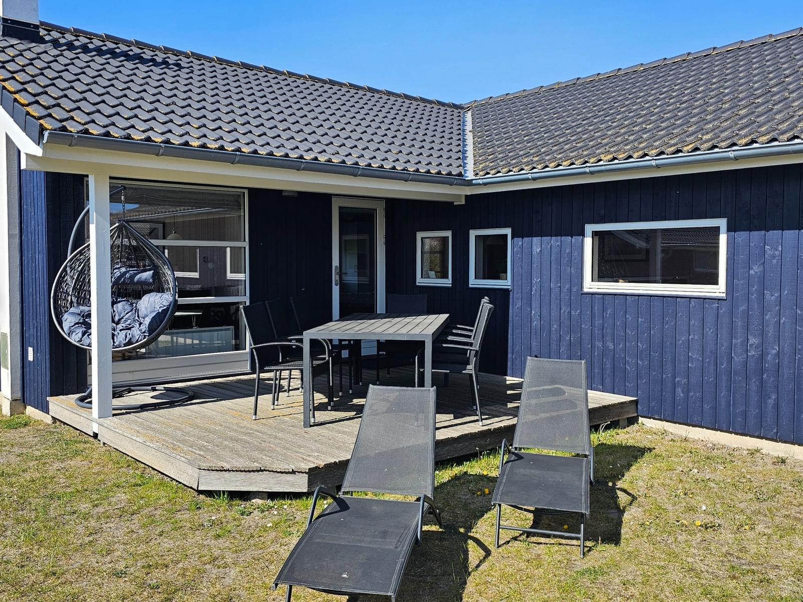 Außenbereich mit Terrasse, Tisch, Stühlen und Liegestühlen vor einem blauen Haus.