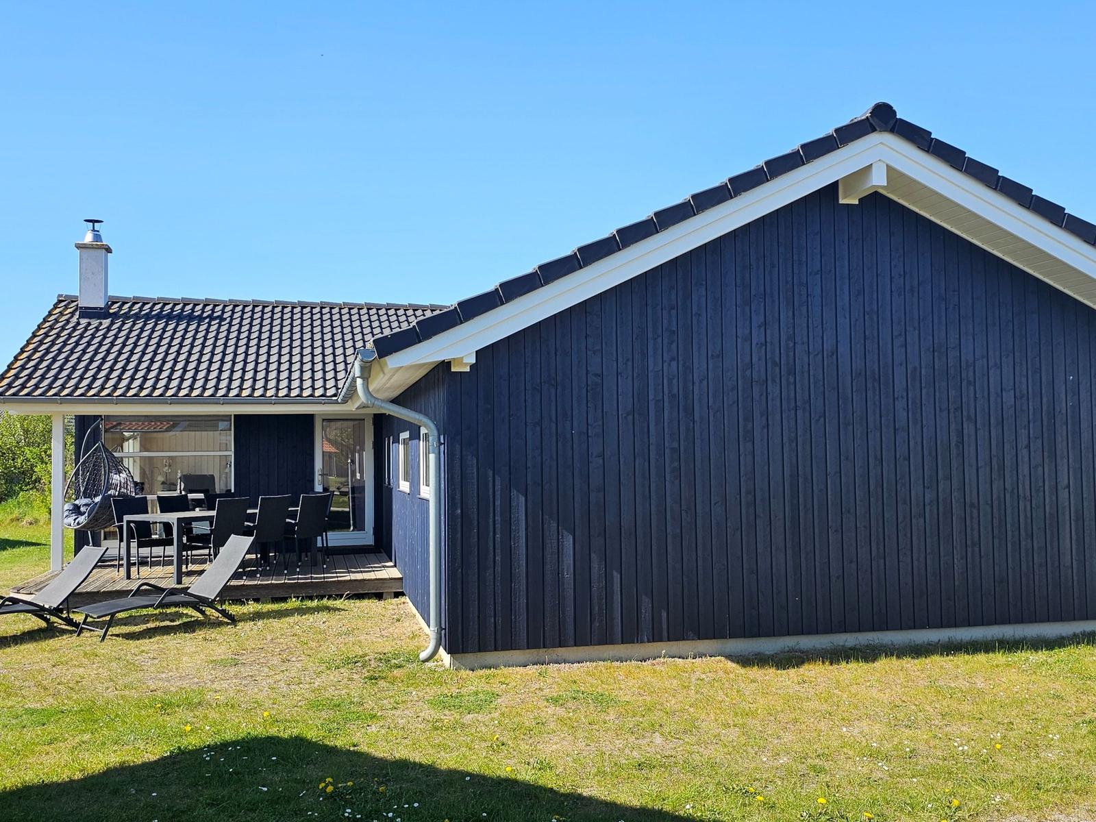 Schwarzes Holzhaus mit Terrasse, Garten und Sonnenliegen unter blauem Himmel.