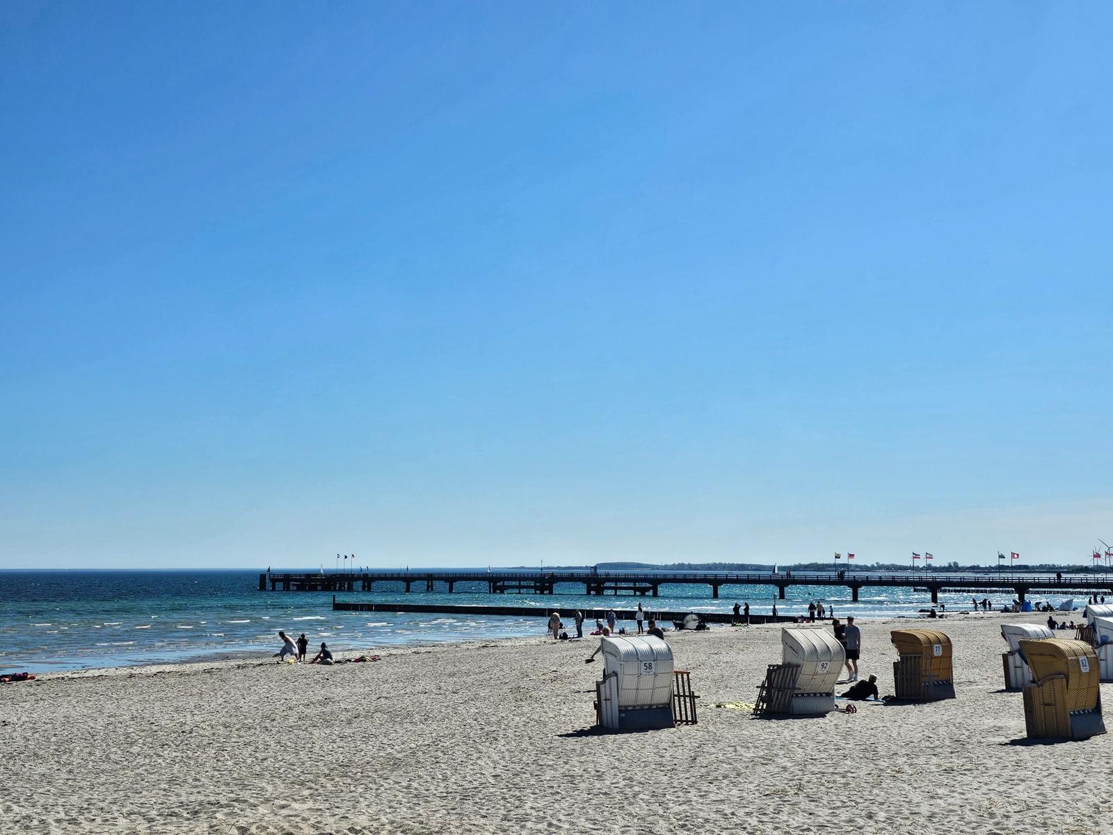 Strand mit Strandkörben und Steg im Hintergrund unter blauem Himmel.
