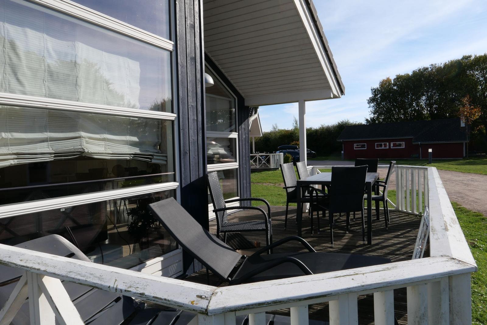 Terrasse mit Tisch, Stühlen und Liegestuhl. Blick auf grünen Garten und rotes Gebäude.