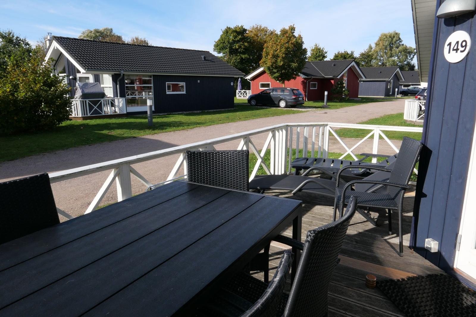 Terrasse mit Tisch und Stühlen. Im Hintergrund sind Ferienhäuser und ein Parkplatz zu sehen.
