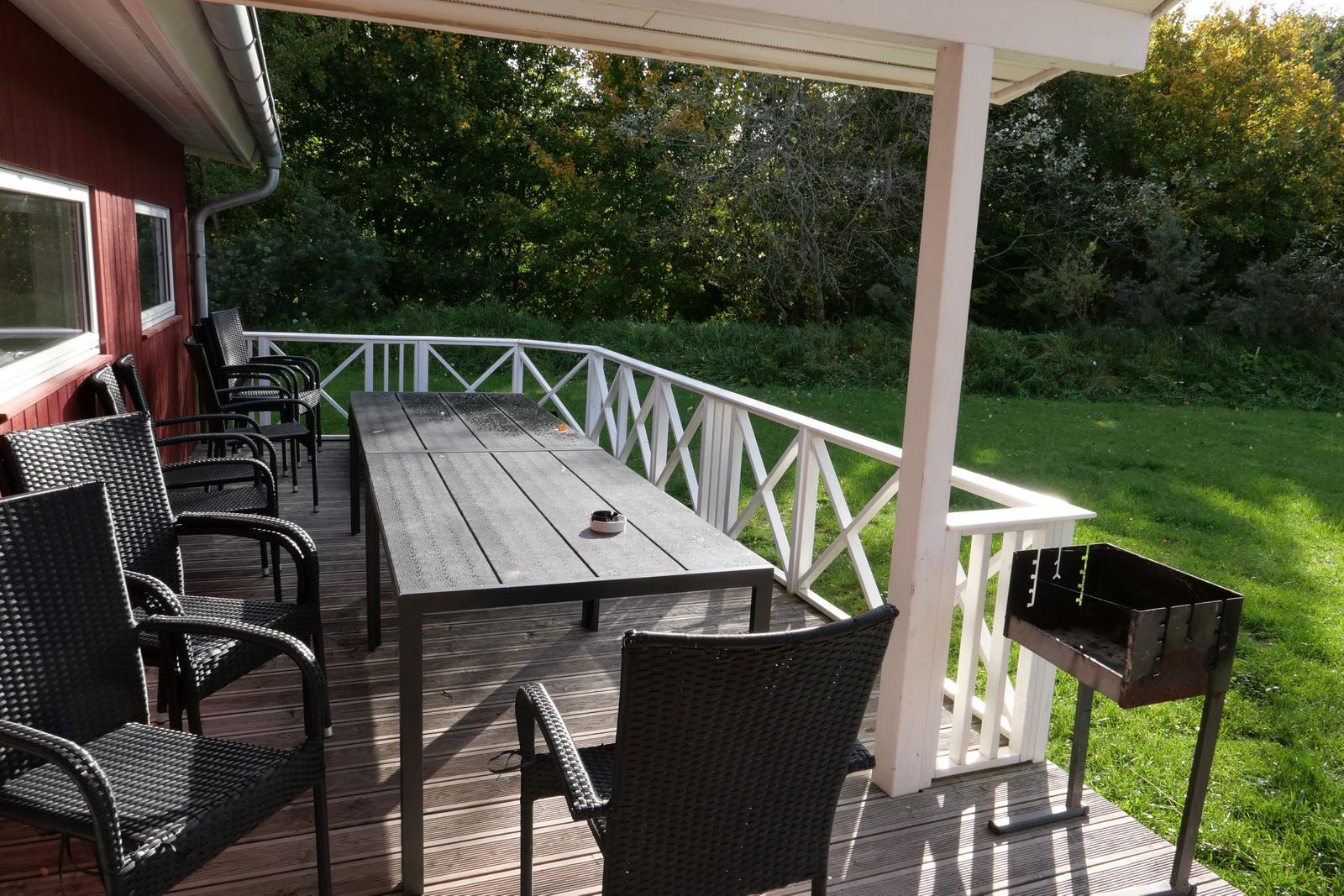 Terrasse mit Tisch, Stühlen und Grill. Hintergrund: grüner Garten und Bäume.