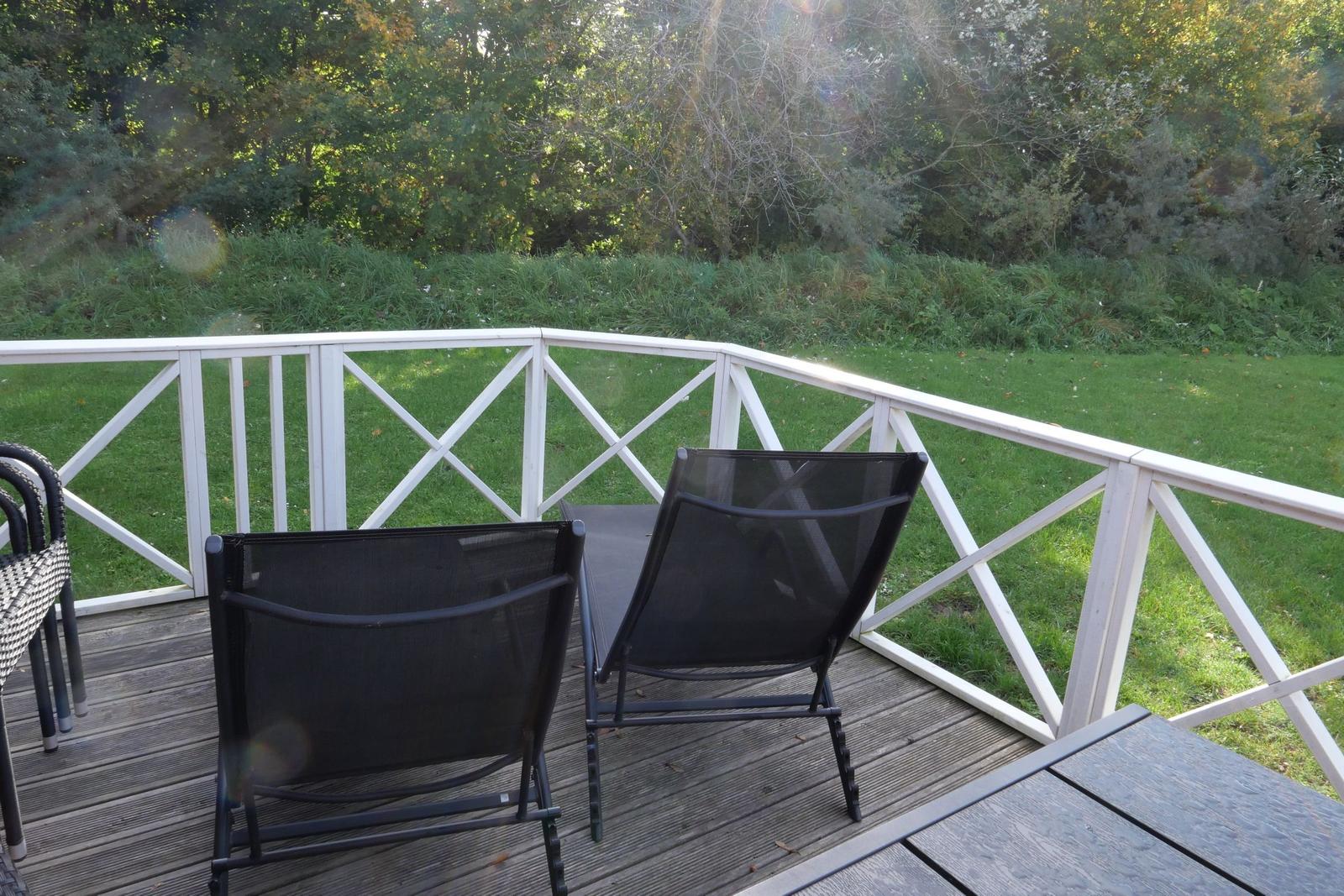 Terrasse mit zwei schwarzen Liegestühlen und weißem Geländer. Grüner Rasen und Wald im Hintergrund.