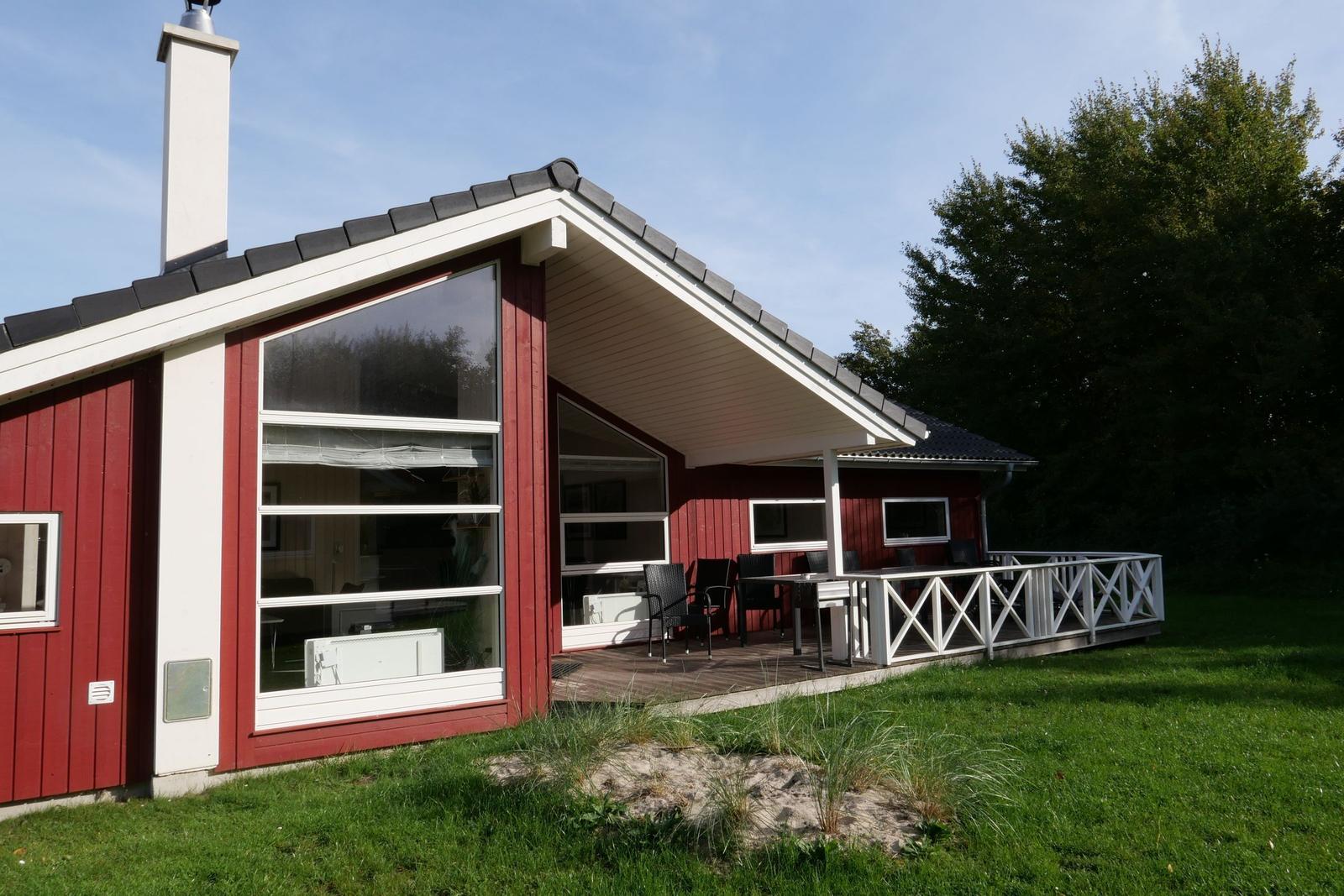 Rotes Holzhaus mit Terrasse und großen Fenstern. Grünflächen und Bäume umgeben das Haus.