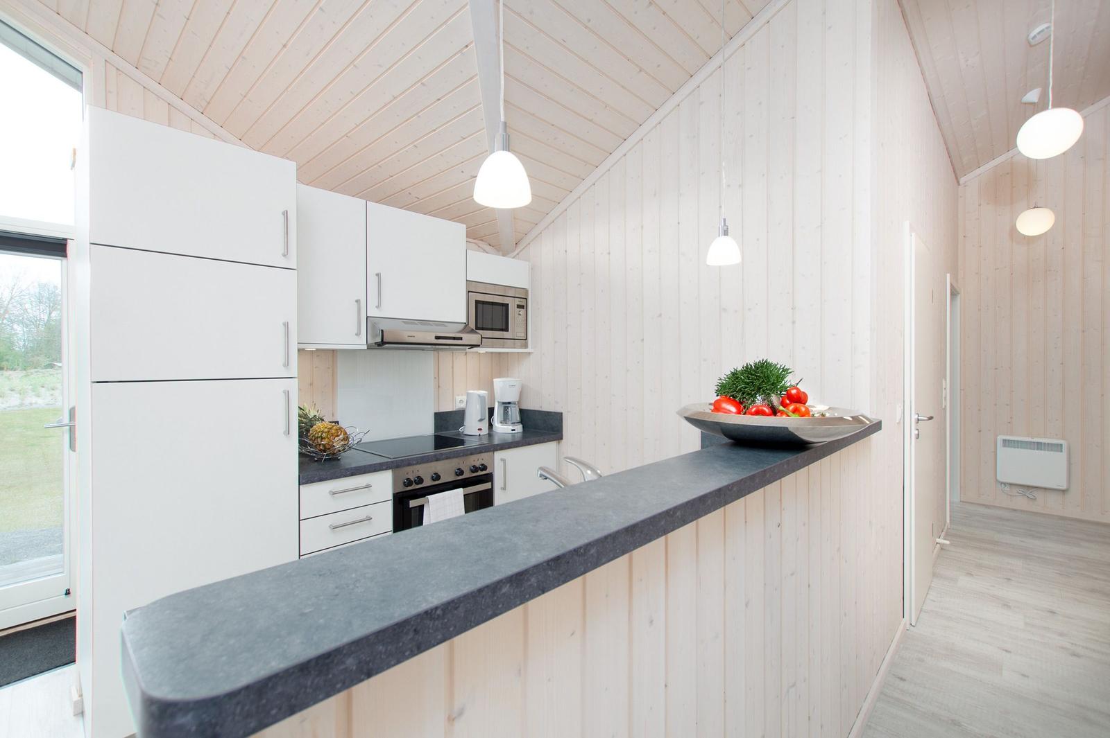 Küche mit weißen Schränken, dunklem Arbeitsplatz und Holzwänden. Auf der Theke steht eine Schale mit Früchten.