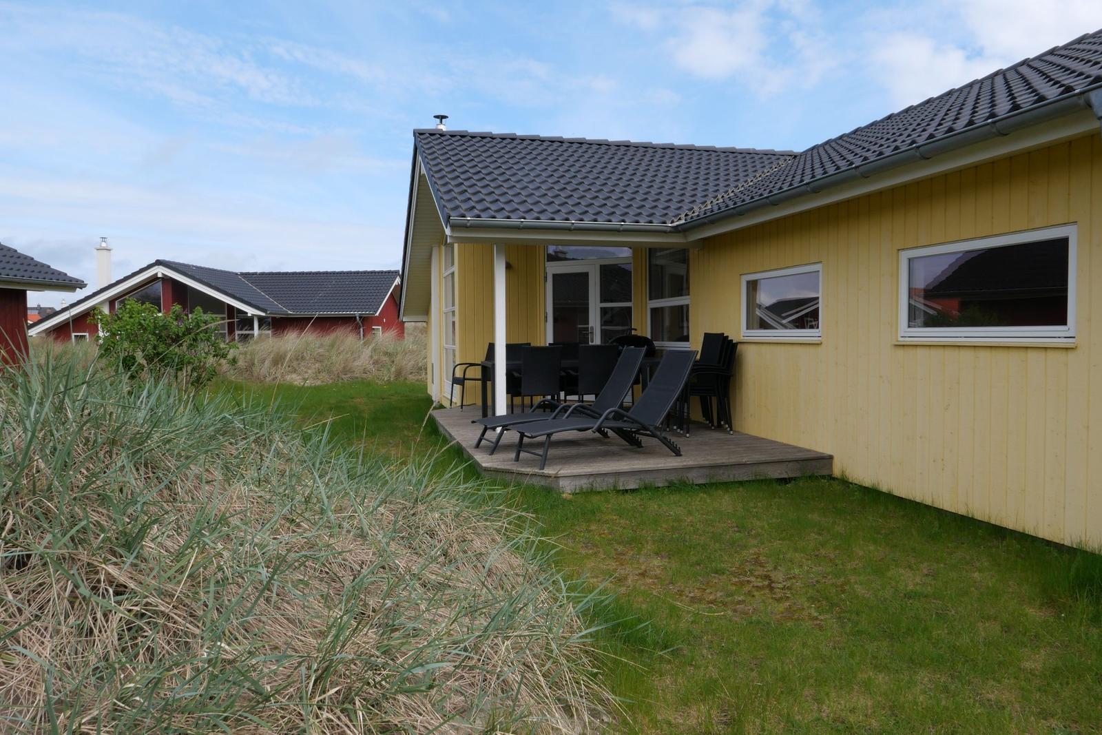 Gelbes Haus mit Terrasse, Liegestühlen und Garten. Im Hintergrund rote Häuser.