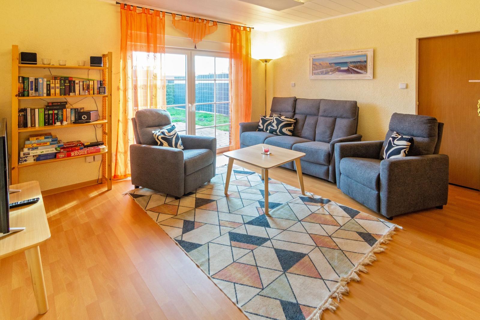 Wohnzimmer mit grauer Couch, zwei Sesseln, Tisch und Teppich. Fenster mit orangefarbenen Vorhängen.
