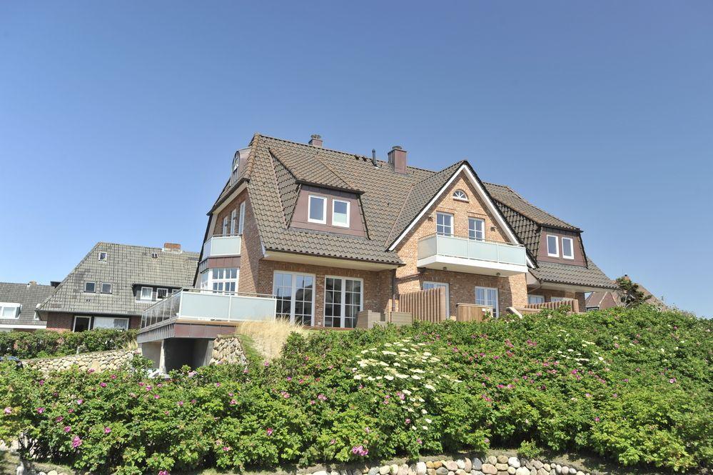 Haus mit Balkon und Garten, umgeben von Blumen und Grünflächen unter blauem Himmel.