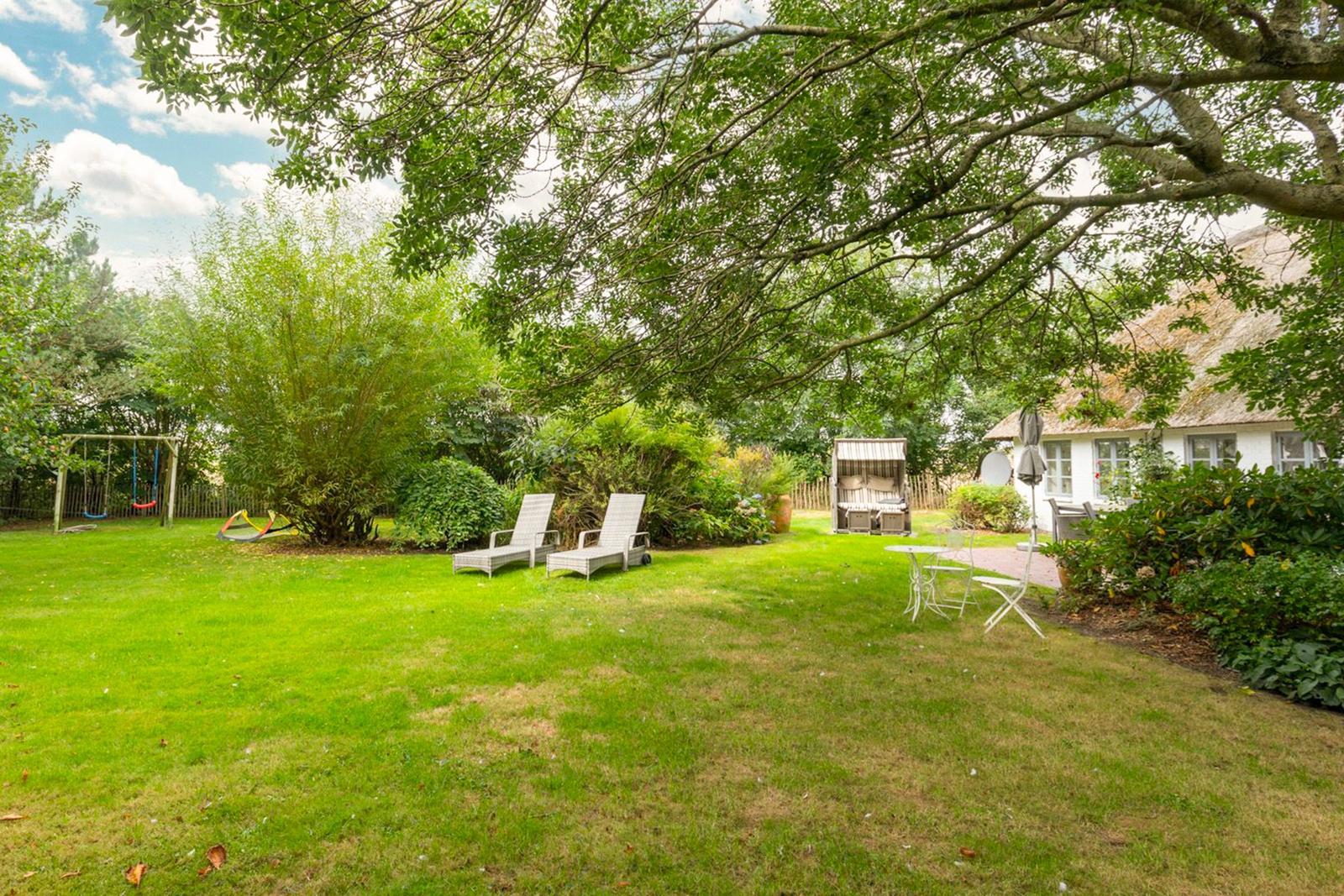 Großer Garten mit Liegen, Tisch, Schaukel und strohgedecktem Haus unter Bäumen.