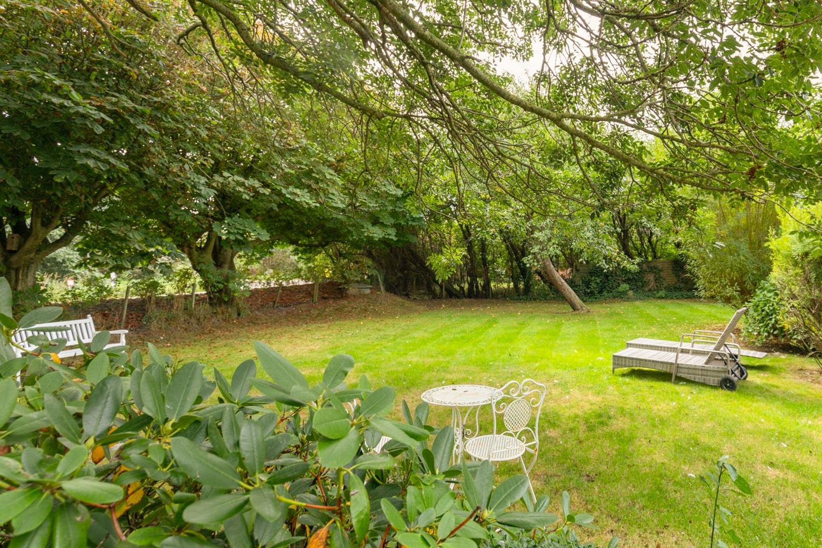 Garten mit grünem Rasen, Bäumen, weißem Tisch und Stühlen sowie Liegestuhl.