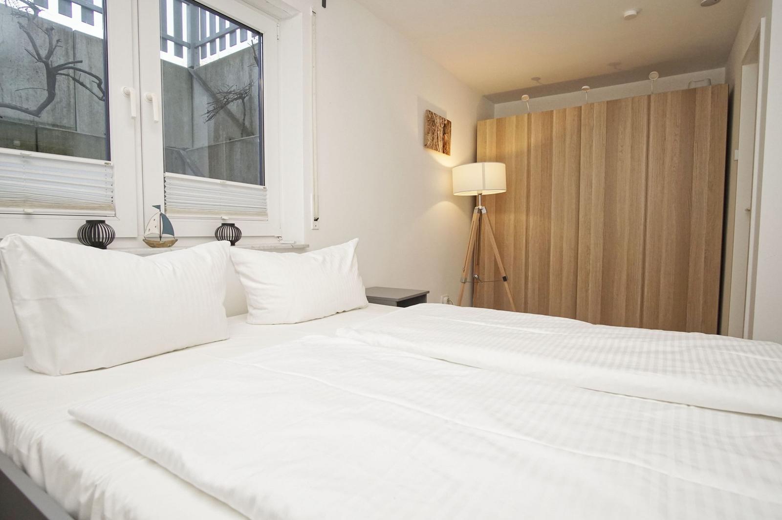 Schlafzimmer mit Bett, Fenster und Holzschrank. Auf dem Fensterbrett stehen Dekorationsgegenstände.