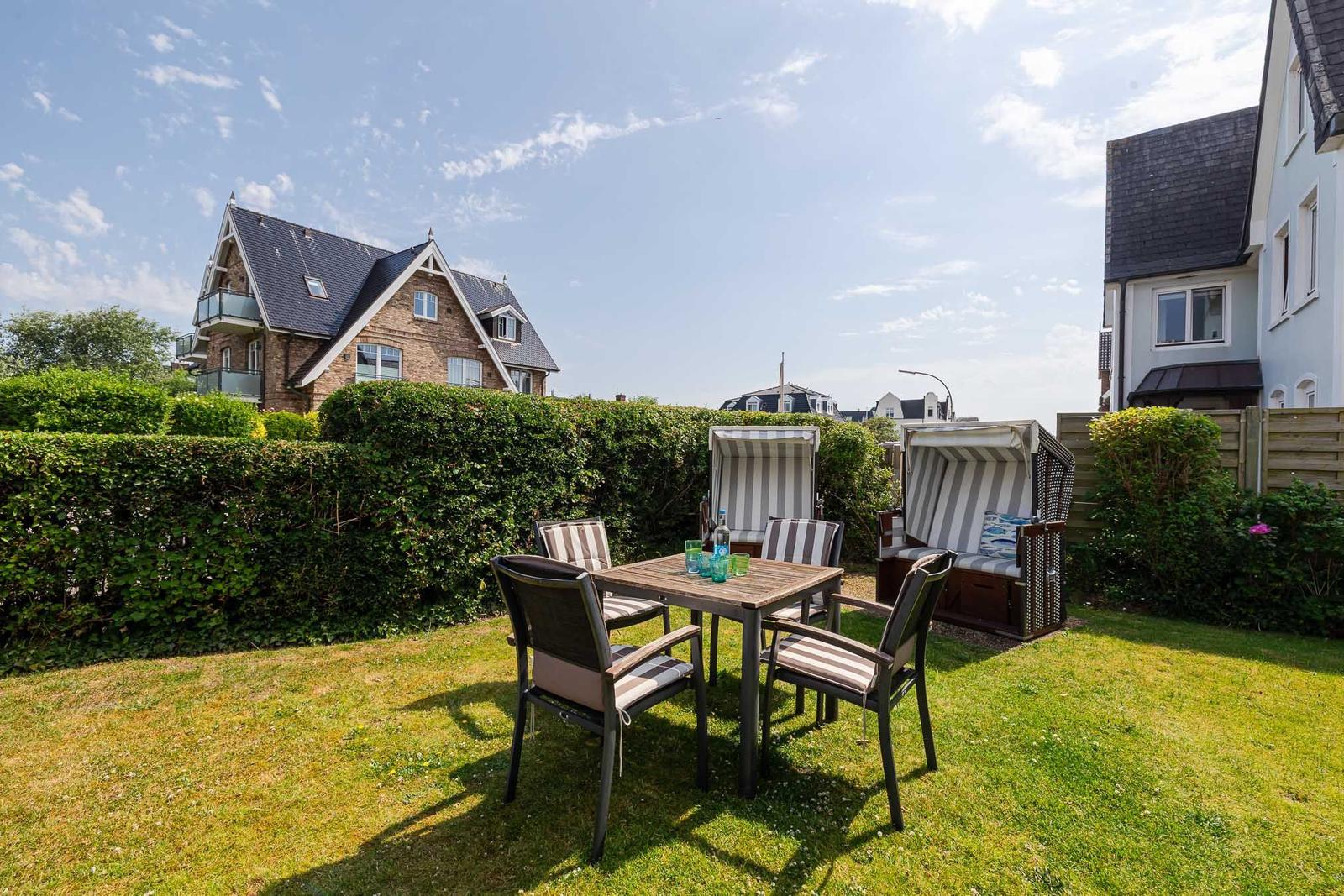Garten mit Tisch, Stühlen und Strandkörben. Hinterhof mit Hecke und Häusern.