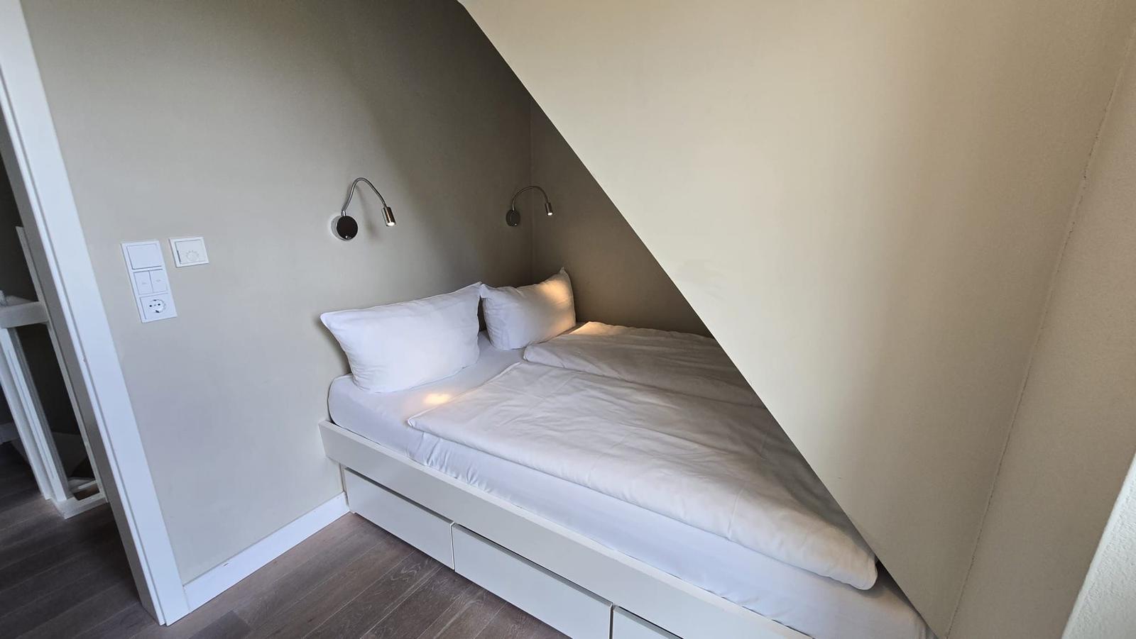 Schlafzimmer mit Doppelbett unter Dachschrägen, Wandlampen und Holzboden.