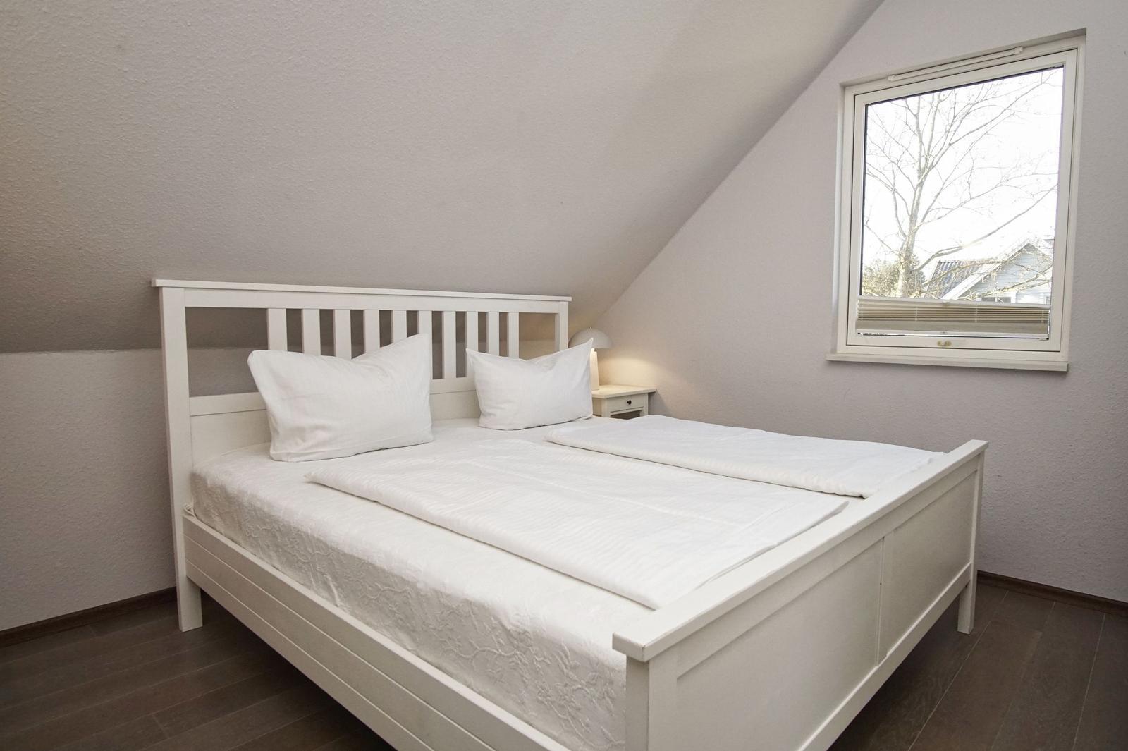 Schlafzimmer mit weißem Bett, Nachttisch und Fenster mit Blick nach draußen.