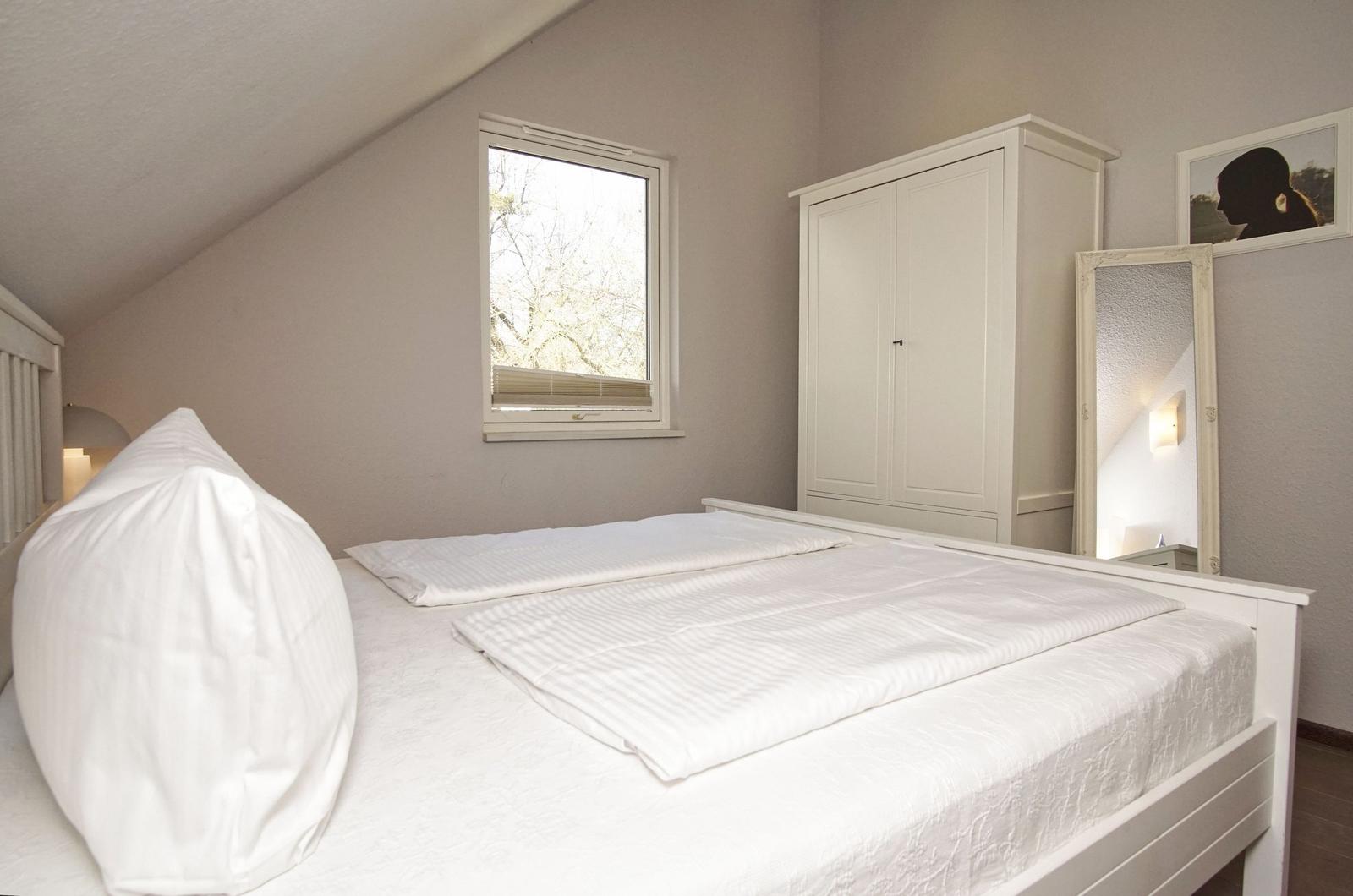 Schlafzimmer mit weißem Bett, Kleiderschrank und Fenster. Spiegel und Wandbild sind sichtbar.