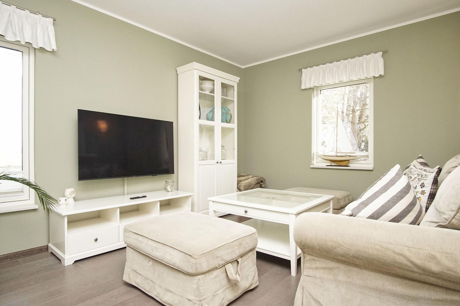 Wohnzimmer mit Sofa, Fernseher und Glas-Tisch. Fenster mit weißen Vorhängen.