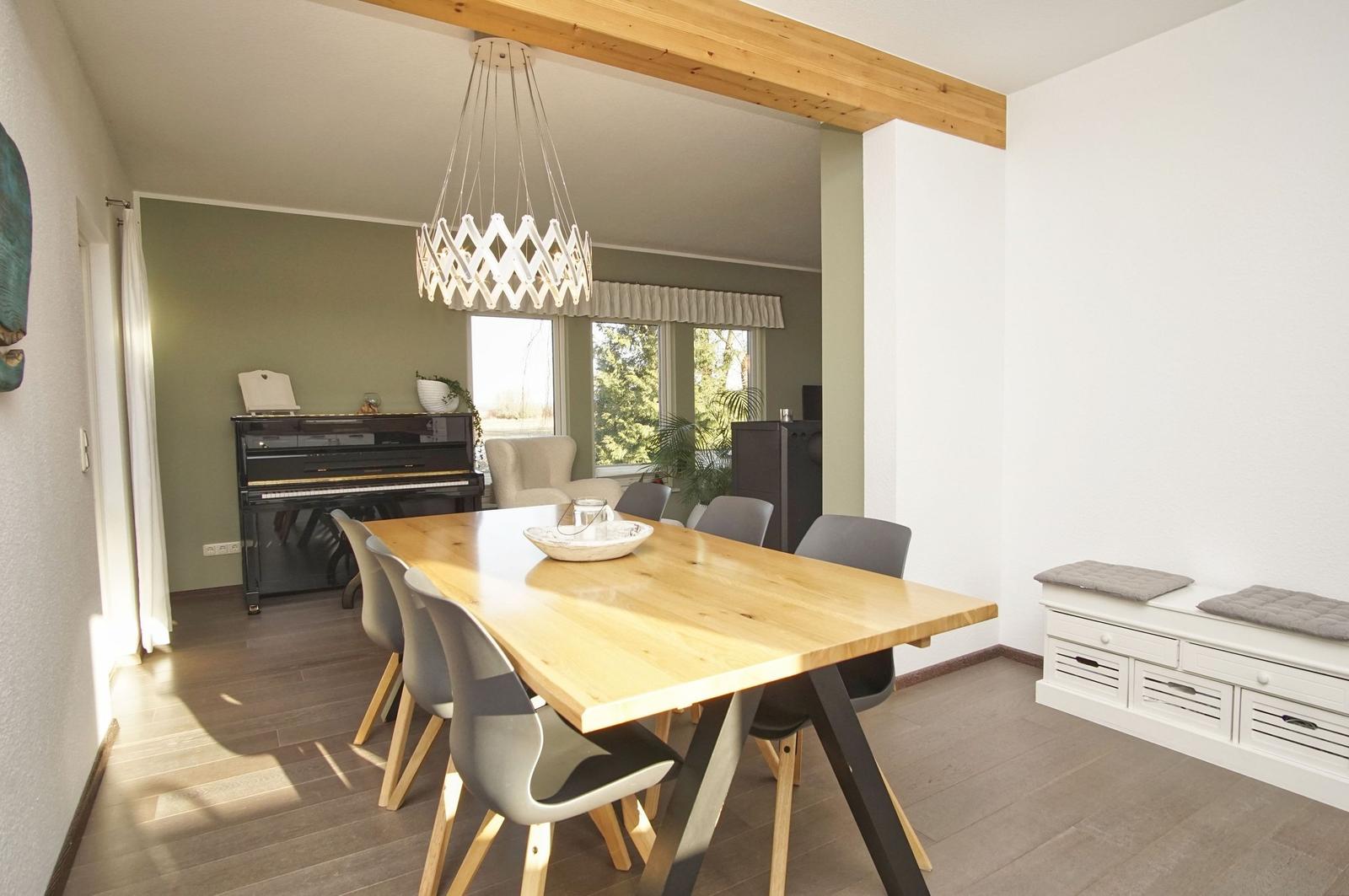 Esszimmer mit Holztisch, Stühlen und Klavier im Hintergrund. Lichtdurchfluteter Raum mit Fensterblick.