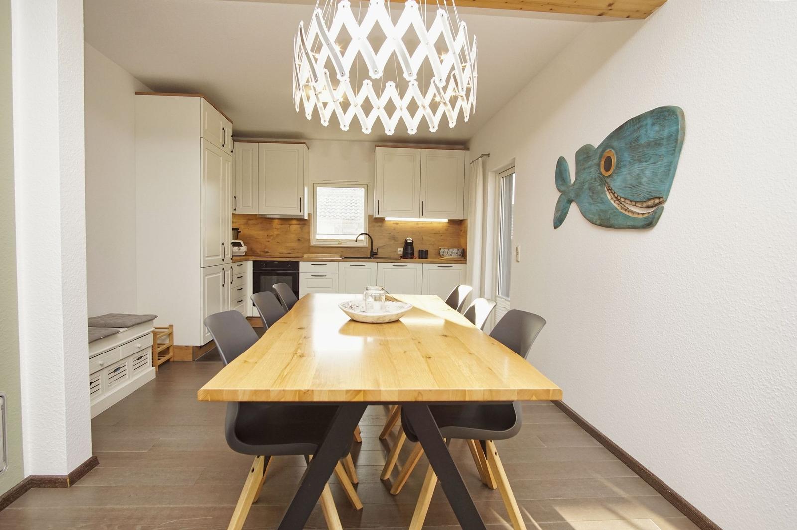 Essbereich mit Holztisch, Stühlen und Küchenbereich im Hintergrund. Wanddekoration mit Fischmotiv.