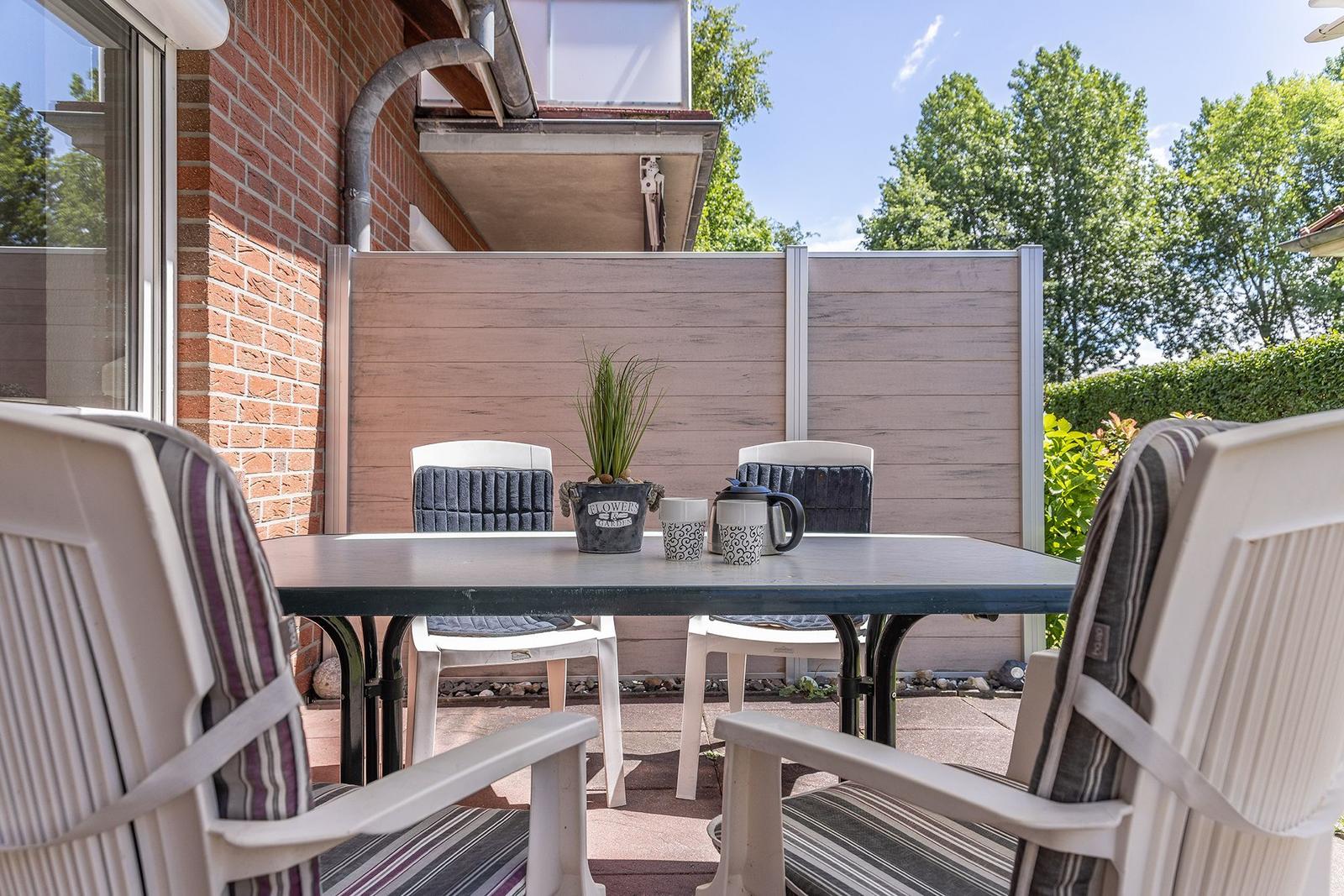 Terrasse mit Tisch und Stühlen, Pflanze und Becher. Hintergrund: Holz-Zaun und Bäume.