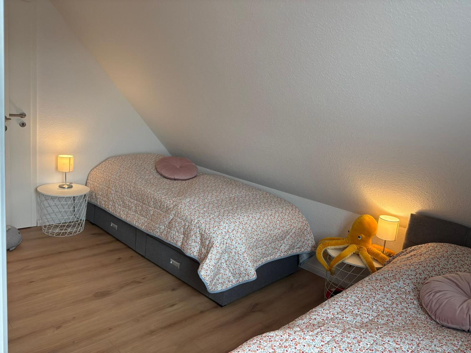 Zwei Betten mit gemusterten Bettwäschen und Nachttischen mit Lampen unter schrägen Wänden.