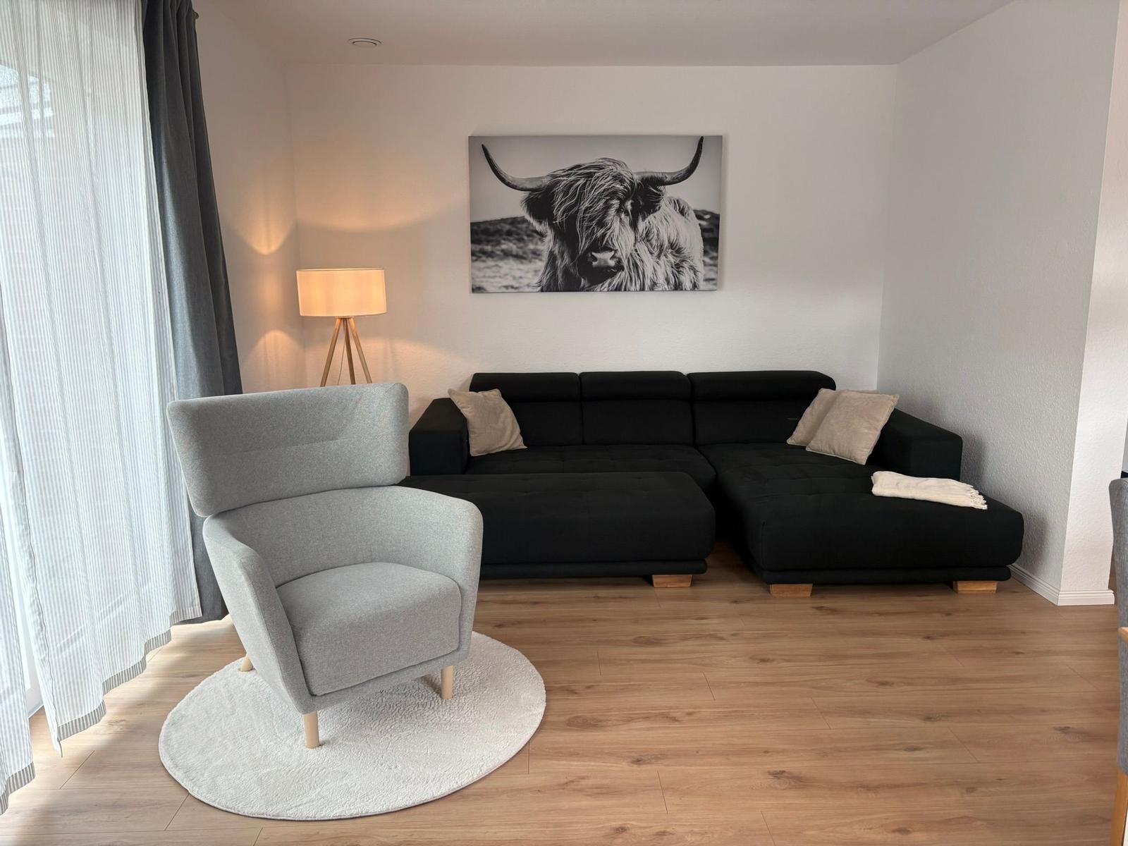 Ein Wohnzimmer mit schwarzem Sofa, grauer Sessel und Wandbild eines Hochlandrindes.