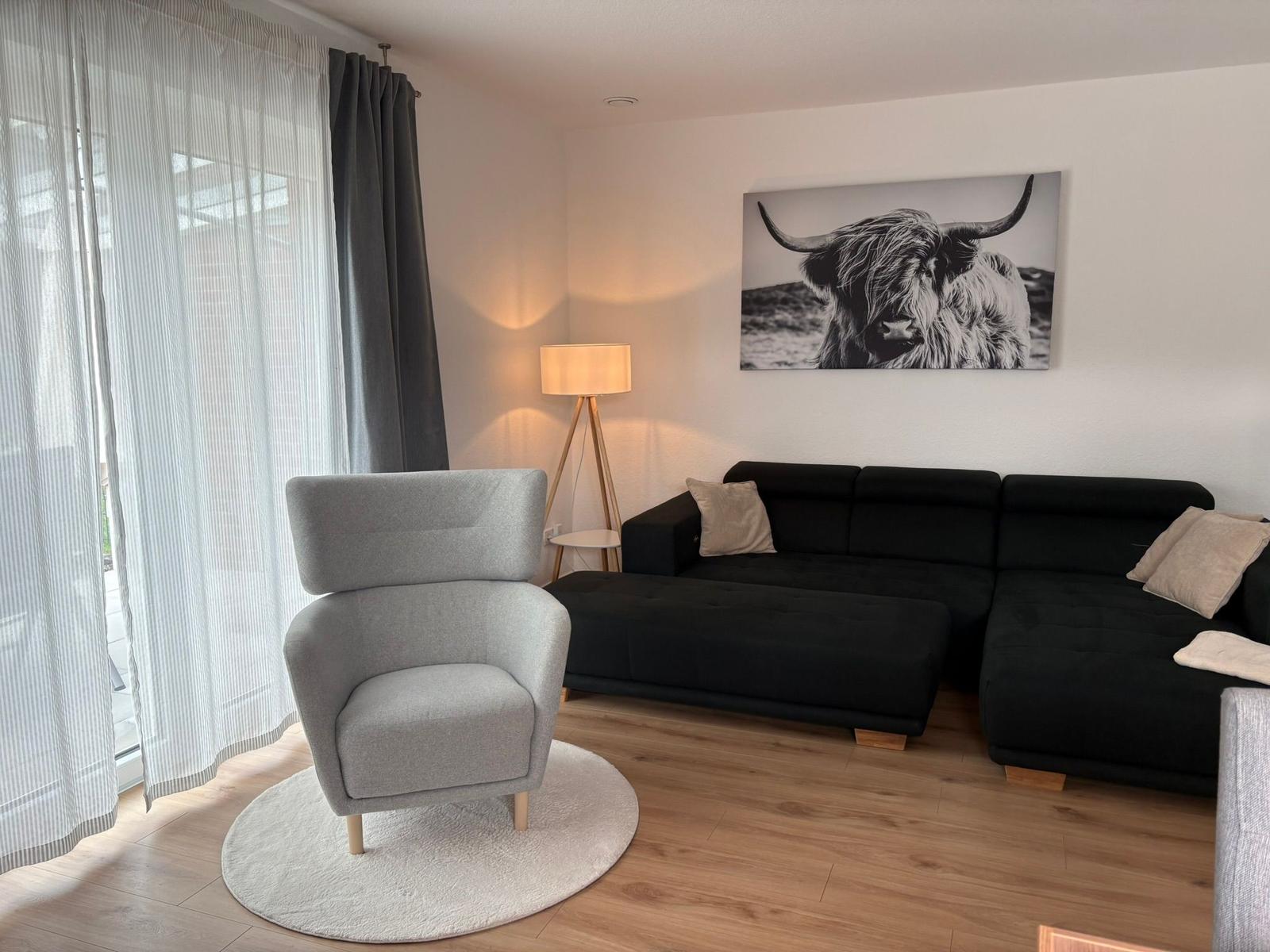 Ein Wohnzimmer mit schwarzem Sofa, grauer Sitzgelegenheit und Wandbild eines Highland-Rindes.