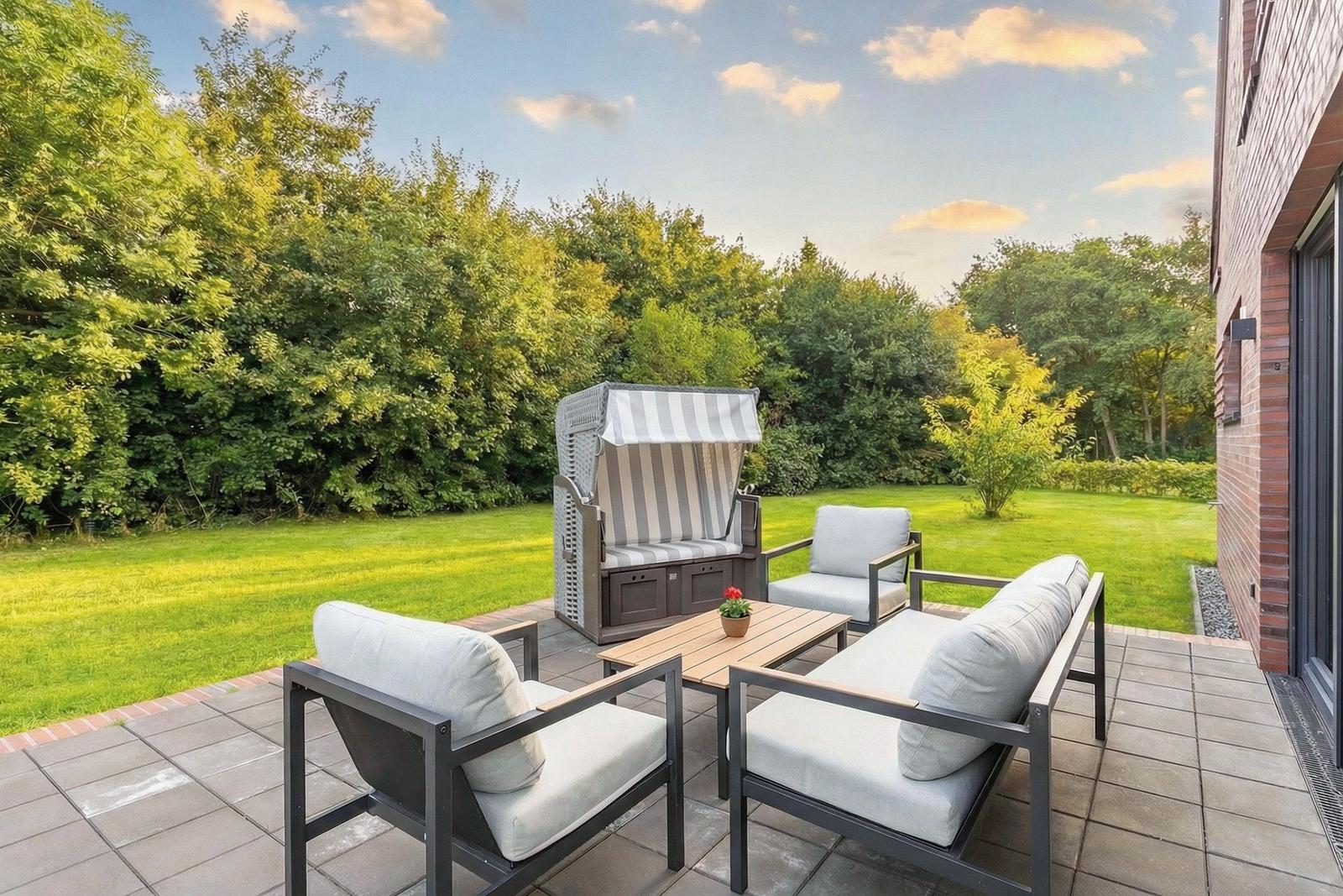 Terrasse mit Gartenmöbeln, Strandkorb und grünem Rasen hinter Bäumen.