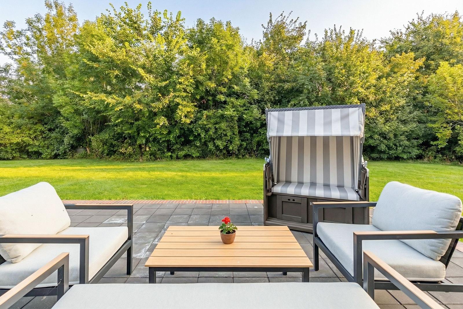 Terrasse mit Sitzgruppe, Strandkorb und Tisch vor grünem Rasen.