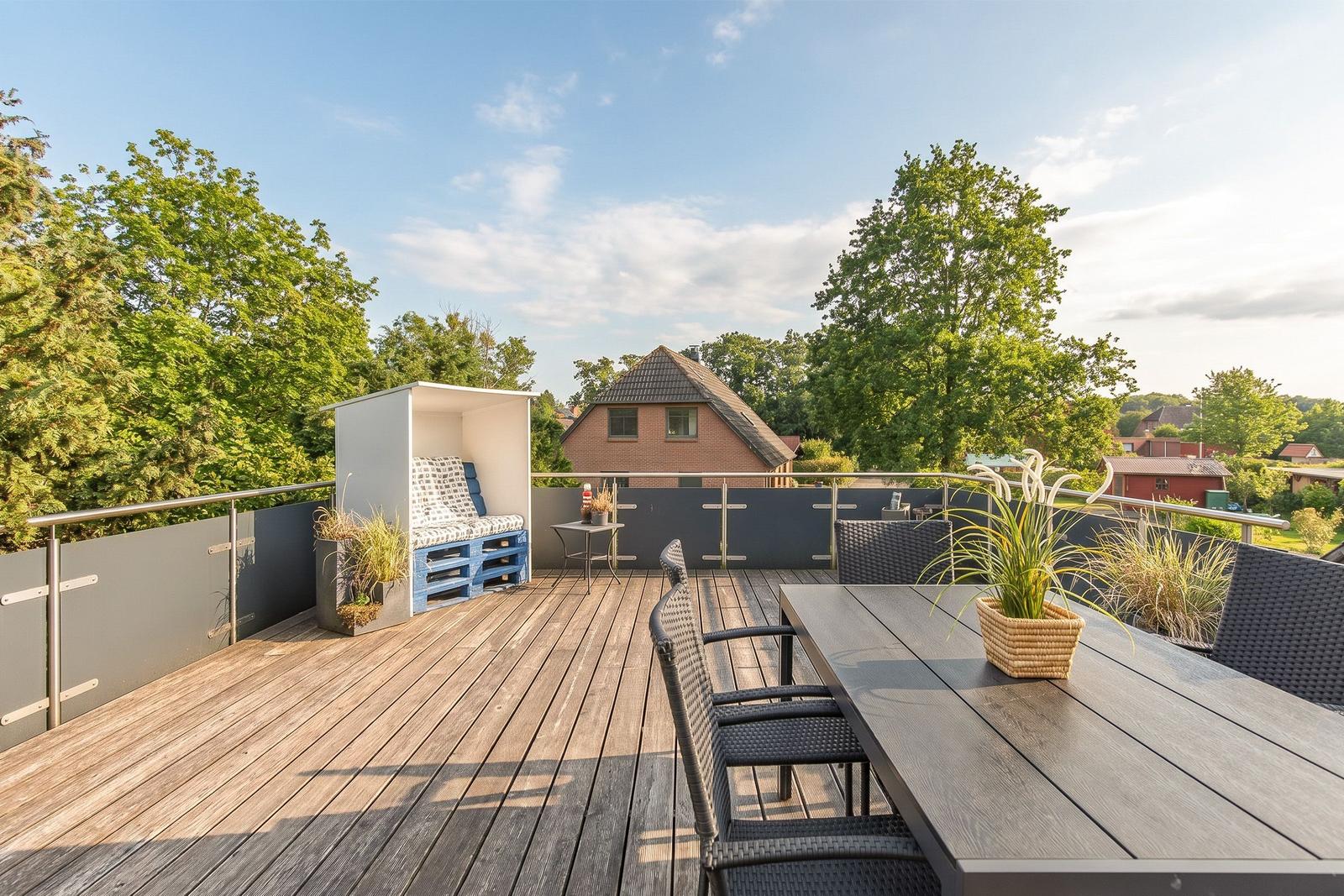 Terrasse mit Sitzgruppe, Tisch und Pflanzen. Blick auf grüne Bäume und ein Haus.