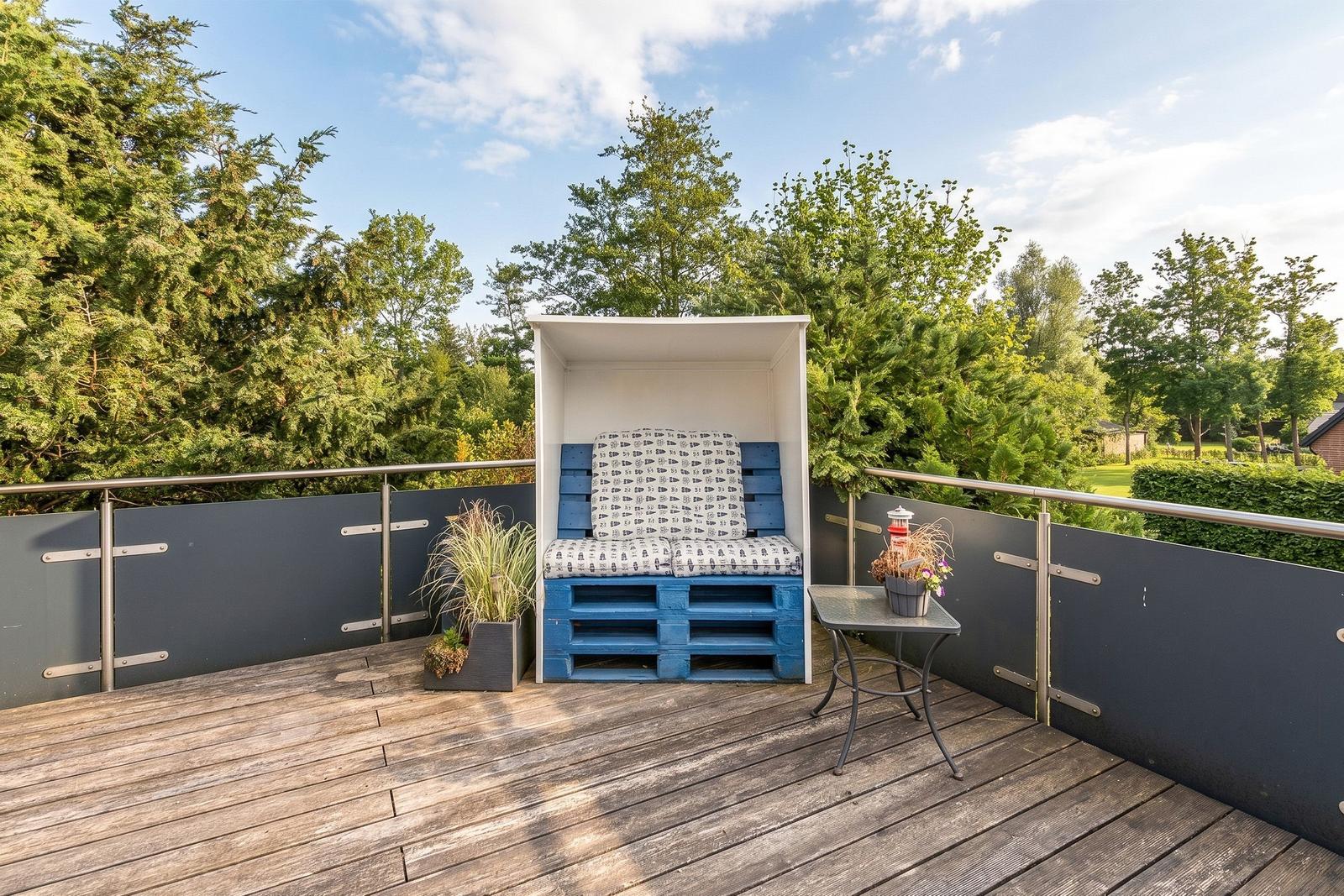 Terrasse mit blauer Sitzbank, Tisch und Pflanzen. Hintergrund: Bäume und Himmel.