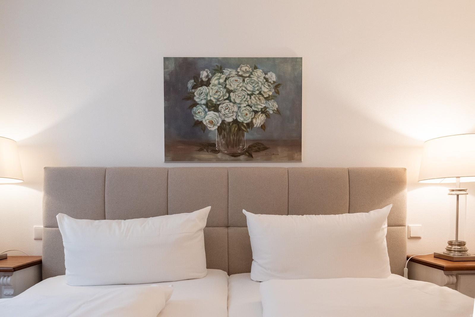 A bed with white pillows and a wooden nightstand. Above the bed hangs a painting with roses.