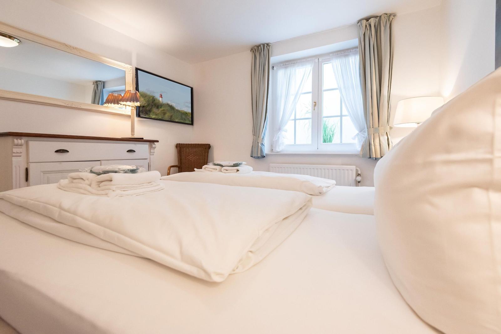 A bedroom with a white bed, window, mirror, and TV.