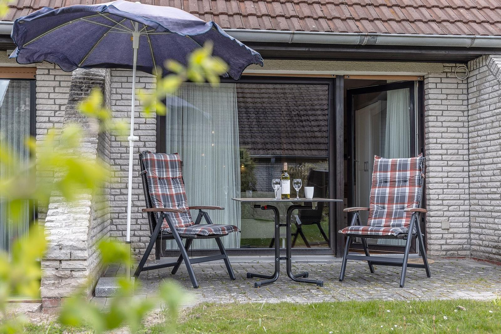Terrasse mit zwei Stühlen, Tisch und Sonnenschirm vor einem Haus mit Glasfront.