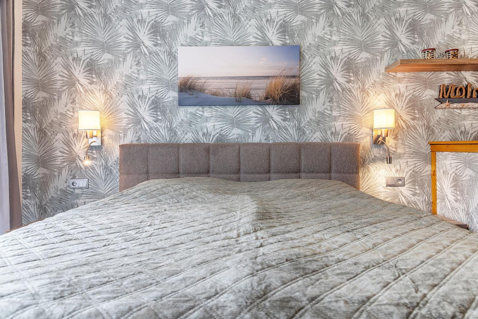 Schlafzimmer mit Bett, Wandtapete und Wandbild. Zwei Wandlampen und Holzregal.